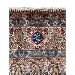 Persian Kirman carpet, the ivory ground field and border decorated with repeating Boteh motifs, interspaced with all-over floral design, blue ground border guards, the top border with signature panel