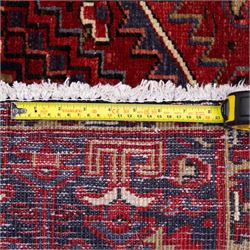 North West Persian Heriz hand-knotted carpet, madder red field with central bold angular steeped medallion with serrated outlines, flanked by stylized palmettes, hooked leaves and geometric motifs, spandrels similarly decorated within a wide geometric border and multiple guard borders in tones of madder red, indigo blue, ivory and olive green, with cotton fringe ends  
