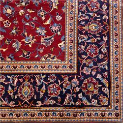 Central Persian Kashan hand-knotted rug, crimson field, decorated with an all-over floral and foliate design incorporating stylized palmettes, rosettes, and scrolling vine work, enclosed by a dark navy blue main border ornamented with flowering branches and palmettes flanked by multiple narrow guard borders with repeating floral motifs, with cotton fringe ends 
