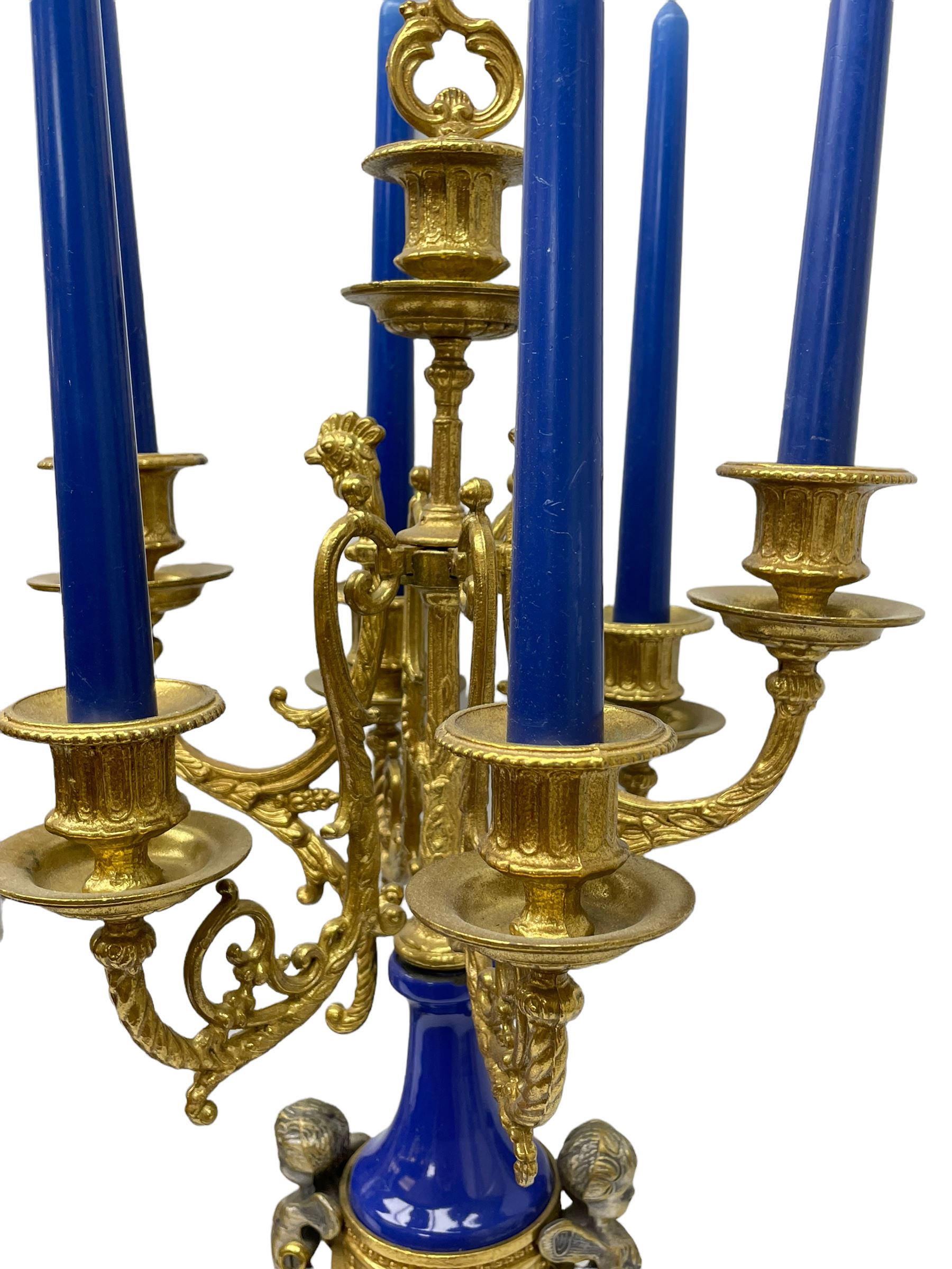 A 20th century continental gilt metal Lyre mantle clock with a pair of matching six light candelabra, spring driven movement housed in a gilt drum case on a brass and cobalt blue base with paw feet, two bronzed metal figures of mythological cherubs with animal legs and cloven hooves supporting a festooned garland swag, eight-day twin barrel striking movement with a floating lever balance escapement, striking the hours and half-hours on two bells, white enamel dial with roman numerals, minute track and pierced steel hands, dial inscribed “Imperial”. 