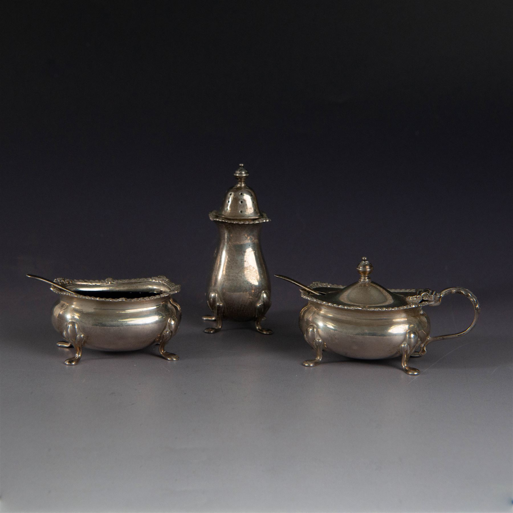 Mid 20th century silver three piece cruet set, comprising pepper shaker, open salt, mustard pot with cover and two condiment spoons, all of oval form with oblique gadrooned rims and upon four paw feet, hallmarked Adie Brothers Ltd, Birmingham 1957, with blue glass liners