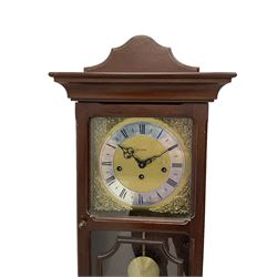 A 20th century wall clock in a simulated mahogany case with a two-part glazed door, brass dial with roman numerals on a silvered chapter ring, brass spandrels, pierced steel hands and visible pendulum, three train Metamec 8-day movement with Westminster chimes sounding on gong rods.

