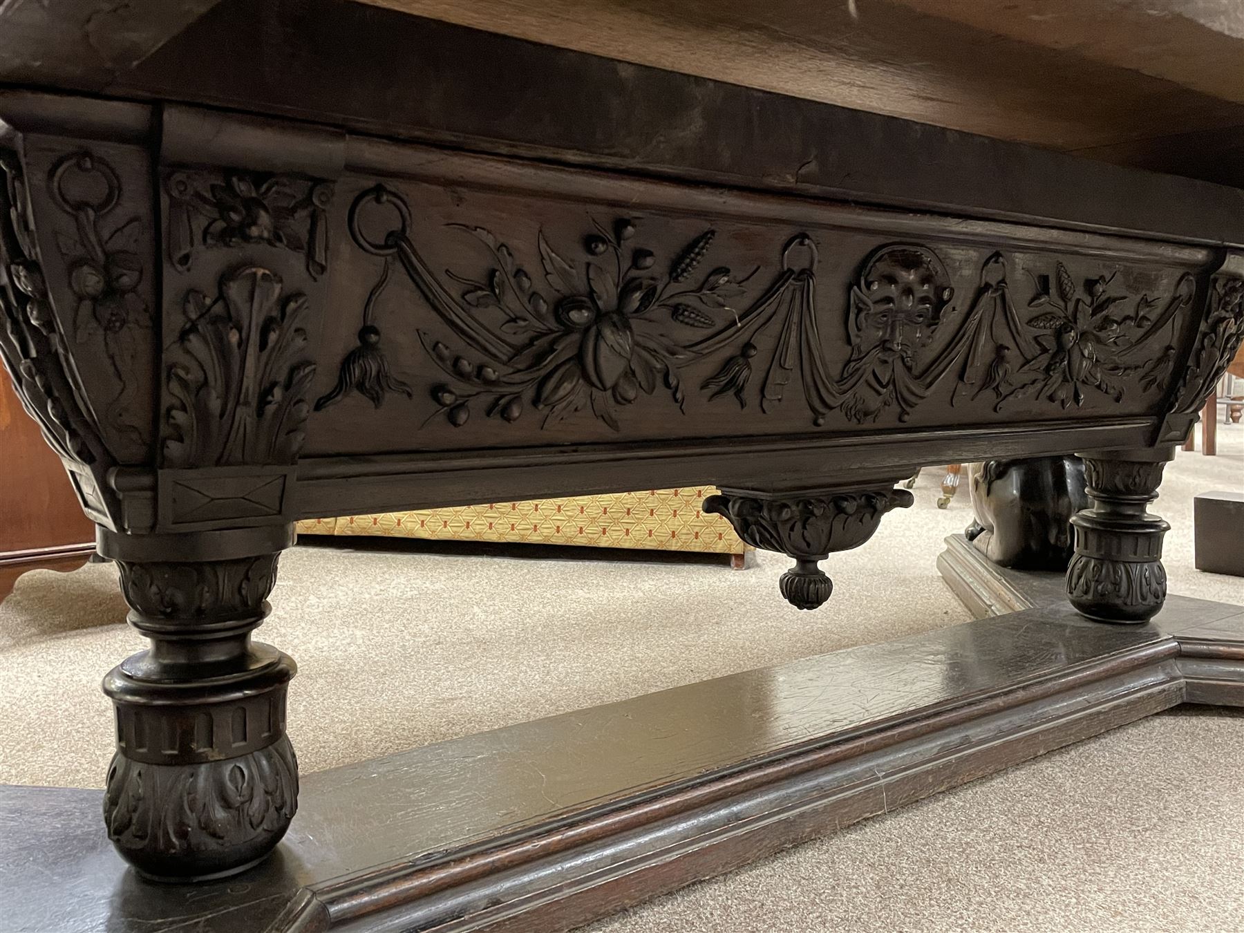 Large 19th century carved oak library table, the rectangular top with lunette carved edge over cartouche panelled and carved  scroll frieze, the central tapering rectangular support relief carved with masks and garlands, on four carved seated lion supports at each corner, on flat angular stretcher with recessed castors stamped ‘Bartholomew & Fletcher, London’
