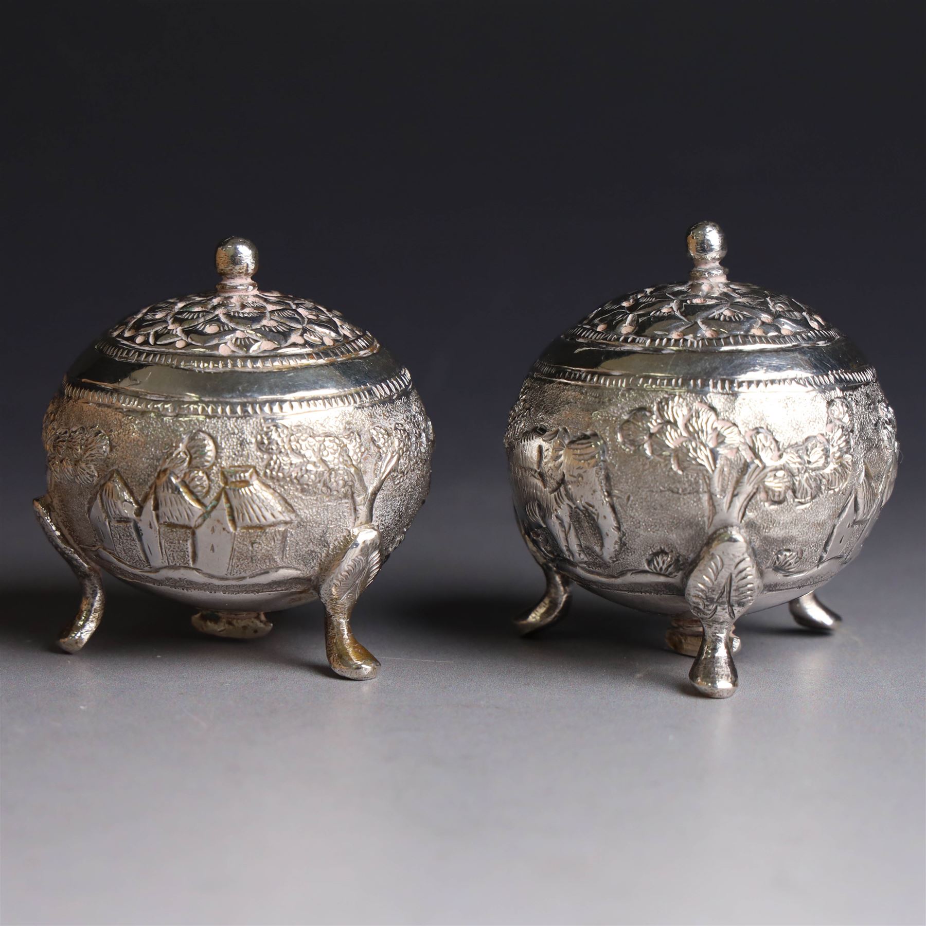 Pair of Middle Eastern silver cruets, of spherical form with pierced tops, embossed with figures, elephants and village scenes, unmarked, together with a pair of silver condiment spoons embossed with floral decoration 
