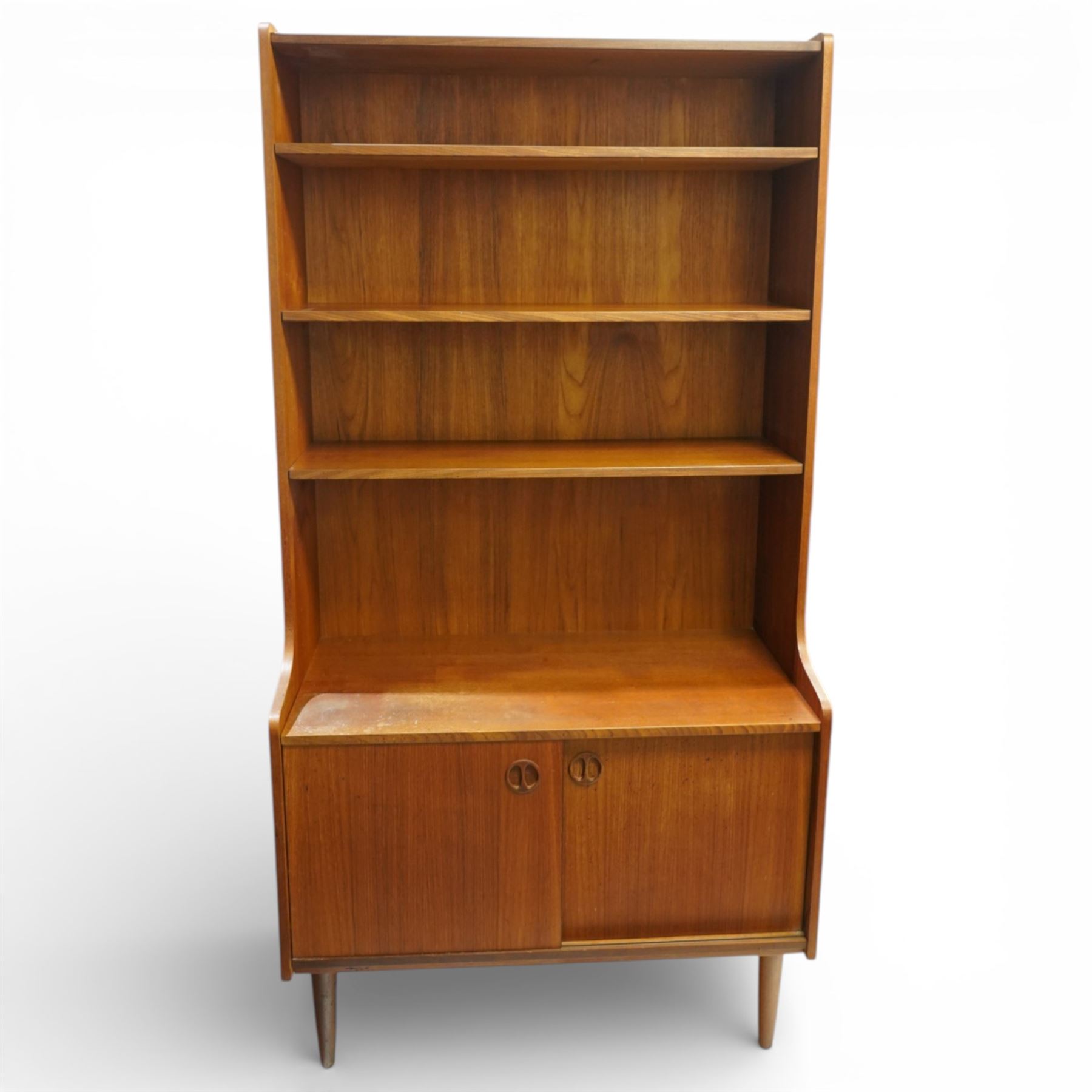 Mid 20th century Scandinavian teak bookcase cabinet in the manner of Johannes Sorth, the upper section with three open shelves over a shaped fall to a lower cupboard enclosed by sliding doors with recessed circular handles, raised on tapering supports