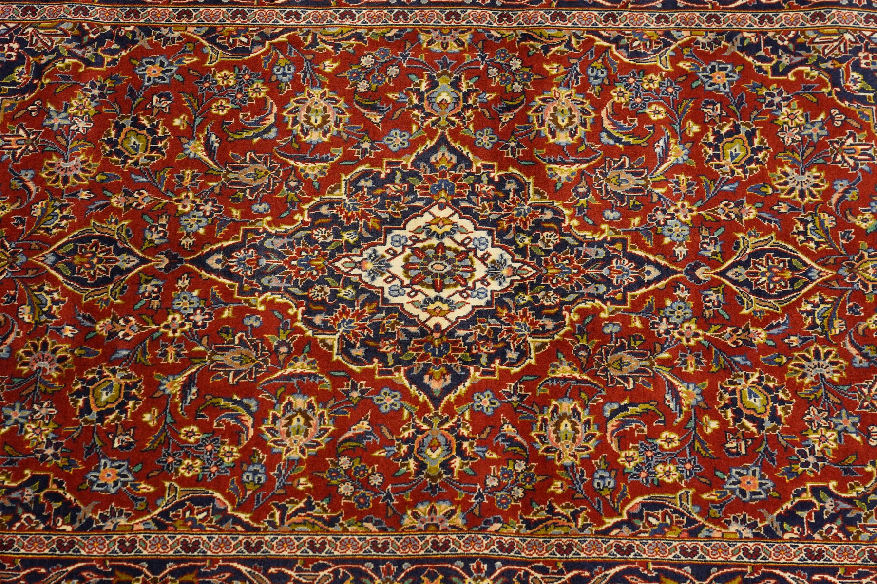 Central Persian Kashan hand-knotted rug, deep red field with central cusped ivory medallion with an all-over arrangement of scrolling floral vines, palmettes and flowering sprays within conforming spandrels, principal navy border with a continuous meandering vine of palmettes and stylized floral motifs, enclosed by multiple narrow guard borders, with cotton fringe ends 