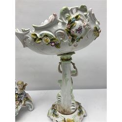 Early 20th century porcelain garniture, the central shaped basket upon a column supported by a female figure, and base with scroll feet, flanked by a pair of four branch candelabra, the scrolling branches and central stem leading to urn shaped sockets over foliate drip pans, upon shaped columns and bases with applied putto, the whole with encrusted flowers throughout, basket H33.5cm, candelabra H52cm