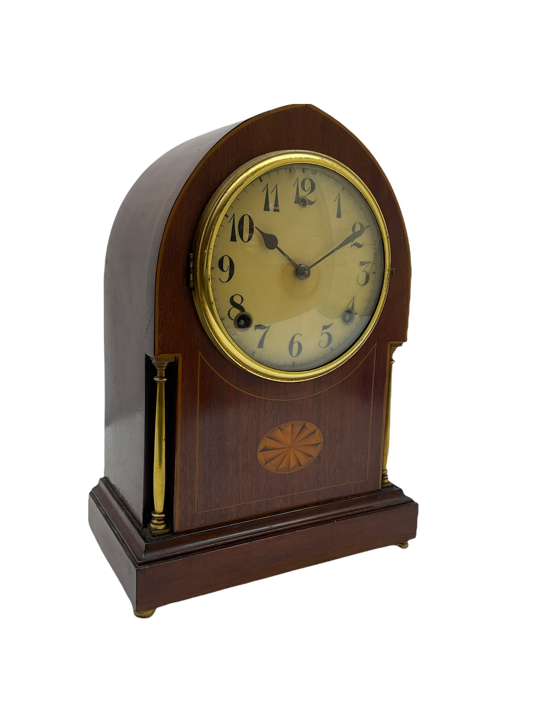 A late 19th century American  mantle clock in a “Lancet” case manufactured by the  Gilbert Clock Factory, Connecticut,  mahogany veneered case with an oval inlay, satinwood stringing to the edge and two recessed brass pillars, on a moulded plinth with four bun feet, enamel dial with upright Arabic numerals and spade hands, fast/slow regulation, with a convex glass and spun brass bezel, eight-day movement striking the hours and half hours on a coiled gong. No pendulum.