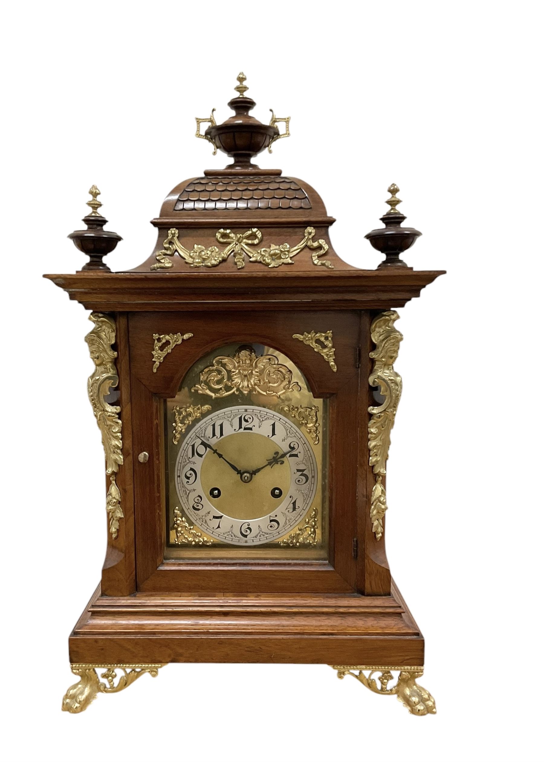 Junghans-German early 20th century walnut cased 8-day mantle clock, with an ogee carved pediment and turned wooden finials, break arch door with brass spandrels beneath flanked by brass caryatids, glazed panels to the sides on a stepped plinth raised on  brass paw feet,  brass dial with a matted centre and silvered chapter with Arabic numerals and Fleur-di Lis steel hands, twin train rack striking movement with a ting tang chime on the quarters. With Key and pendulum. 