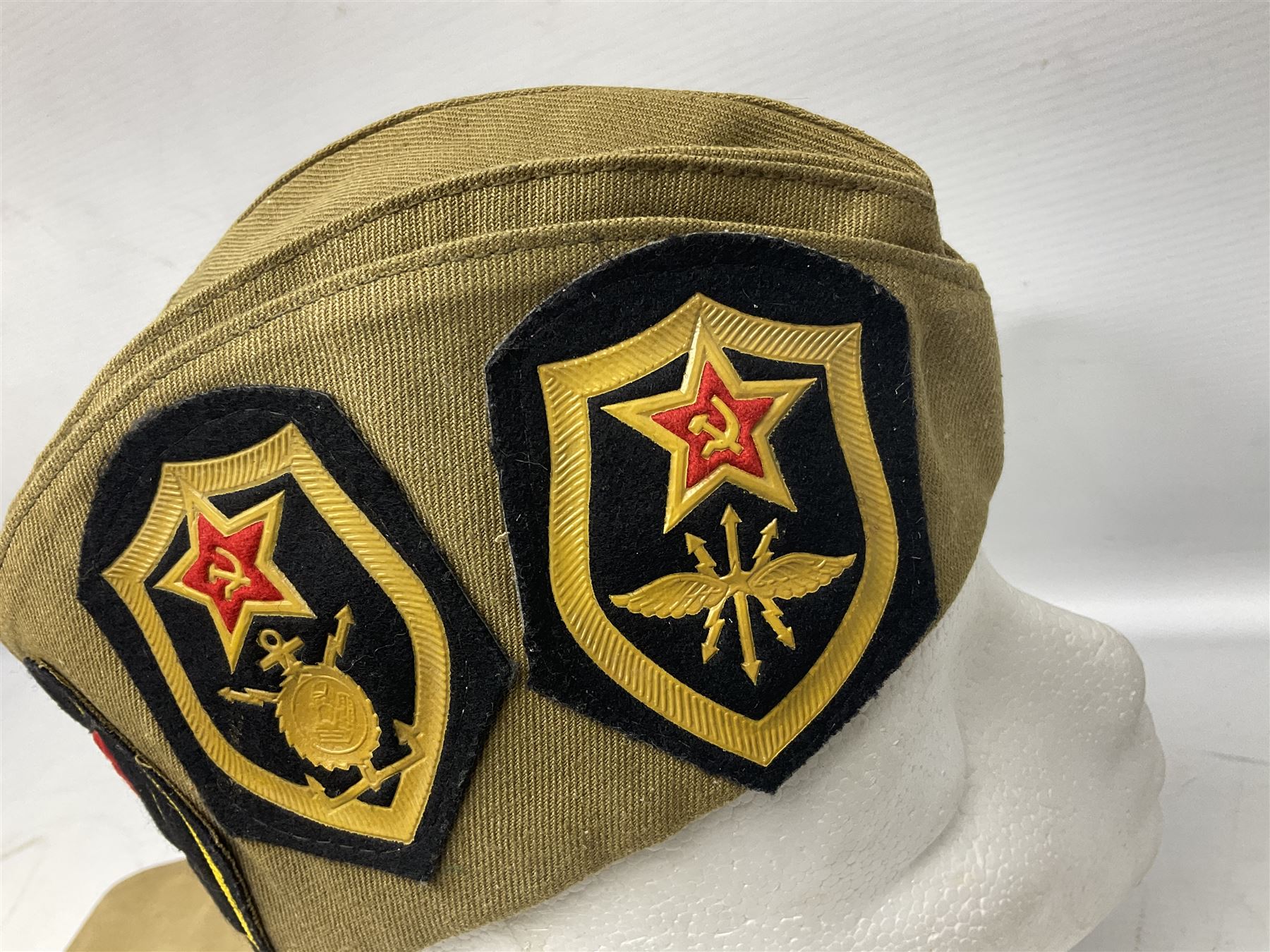 Four side caps - WW2 Russian, post-WW2 Russian and two East German; all with badges, one with multiple metal badges (4)