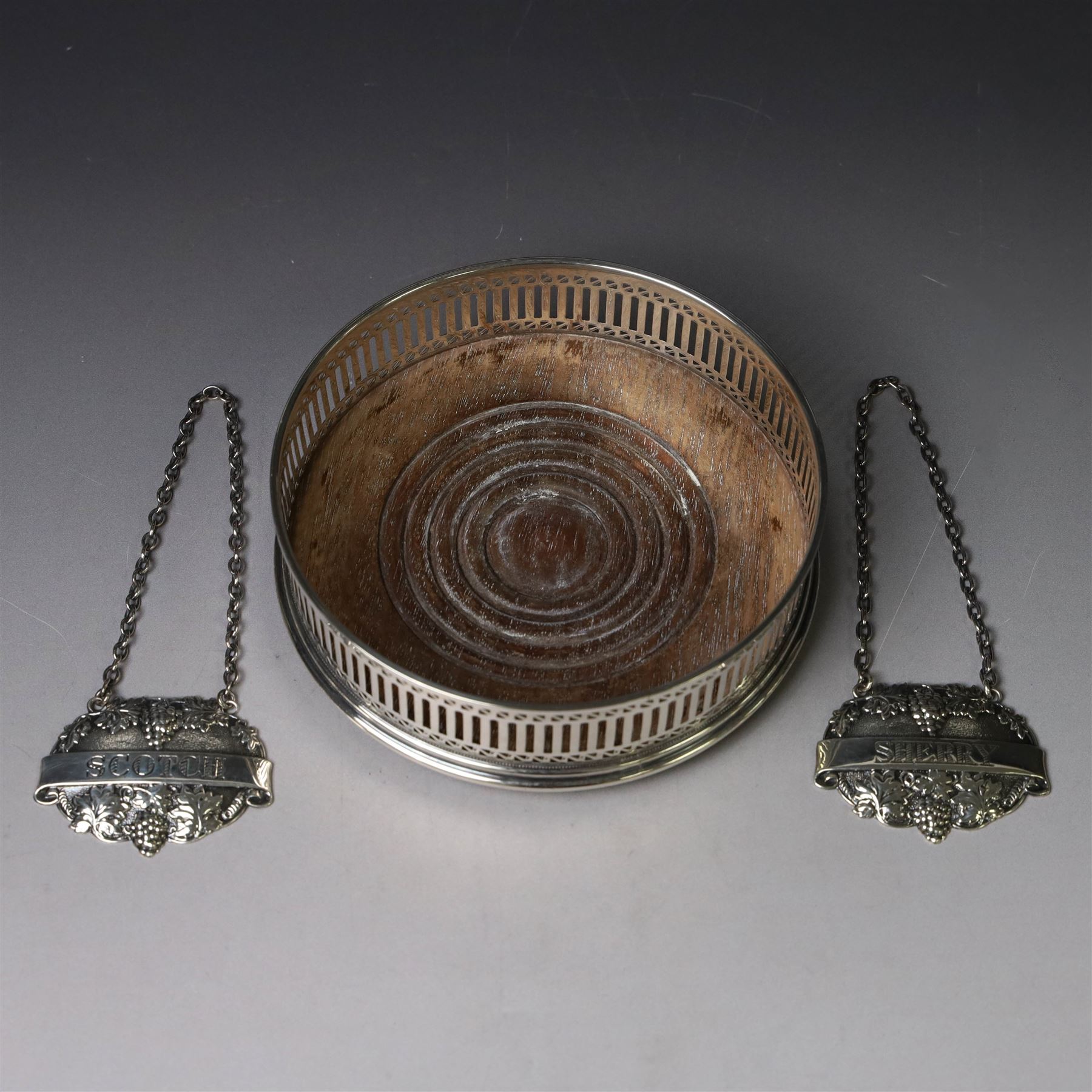 Silver bottle coaster, of circular form with turned wooden centre and pierced sides, hallmarked LGS, London 1977, together with two decanter labels, each with embossed grape vine decoration engraved Scotch and Sherry, hallmarked 