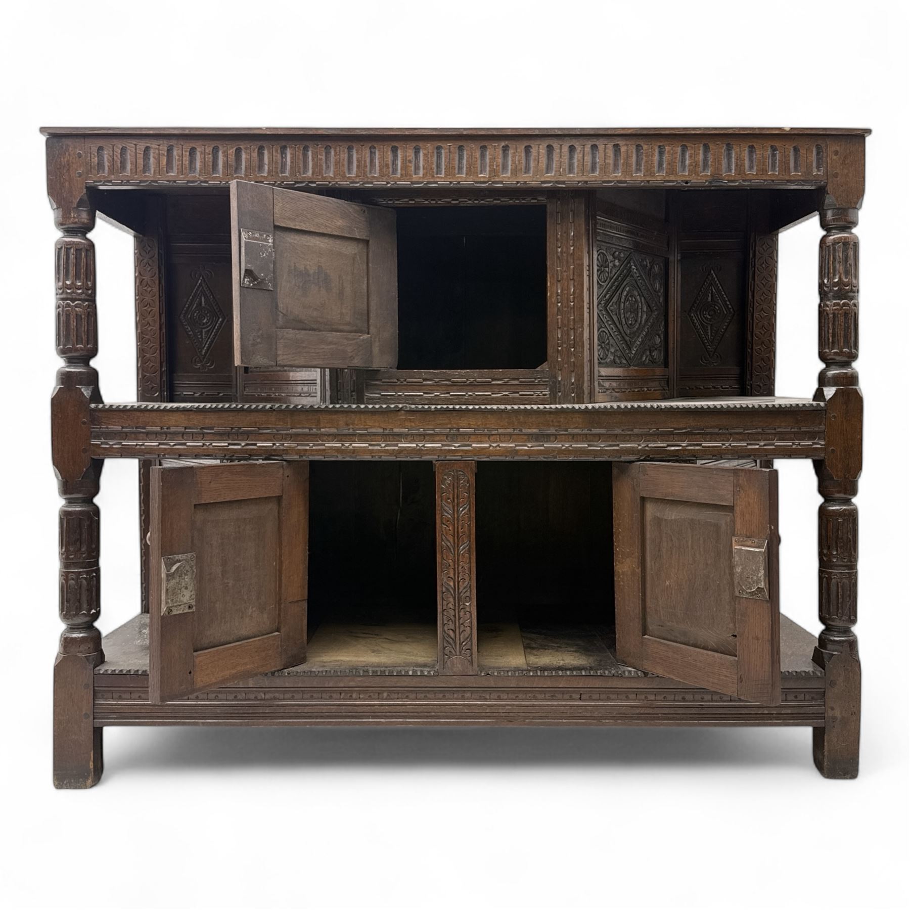 Early 20th-century  oak Jacobean two-tier revival court cupboard, rectangular top with carved notch and stylized foliate carved edge above a carved lozenge pattern frieze and a central cupboard with carved geometric designs and geometric carved panelled sides, above a carved panelled cupboard with two doors of similar geometric design, the whole flanked by two sets of stop-fluted baluster turned columns and supported on stile feet