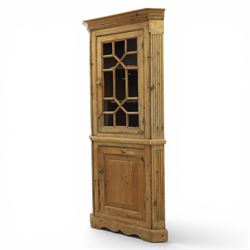 19th century stripped pine corner cabinet, projecting cornice flanked by fluted sides, astragal glazed upper door with brass knob, lower section with fielded panelled door, raised on a shaped plinth base 