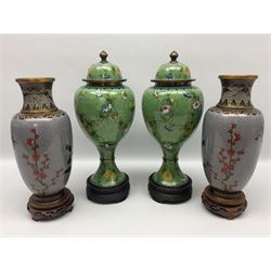 Pair of cloisonné lidded vases of baluster form decorated with blooming branches of flowers upon patterned green ground, together with further pair of cloisonne vases decorated with birds and butterflies amongst peonies and cherry blossom upon patterned lilac ground, all with wood bases, tallest H31cm