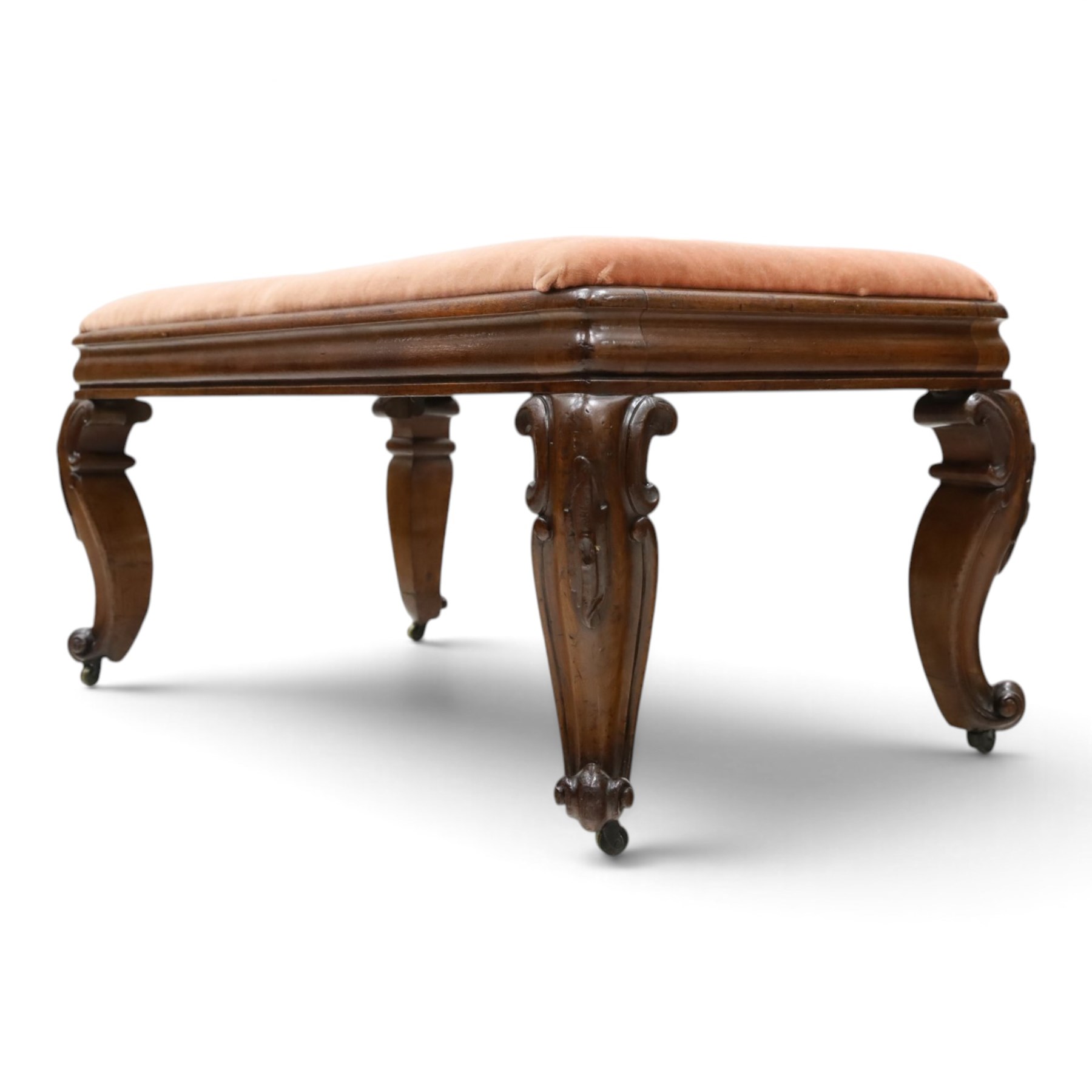 Victorian carved mahogany fender stool of generous proportions, the wide rectangular seat upholstered in rose fabric, shaped frieze rail over C-scroll and cartouche carved cabriole supports with scroll feet and brass castors