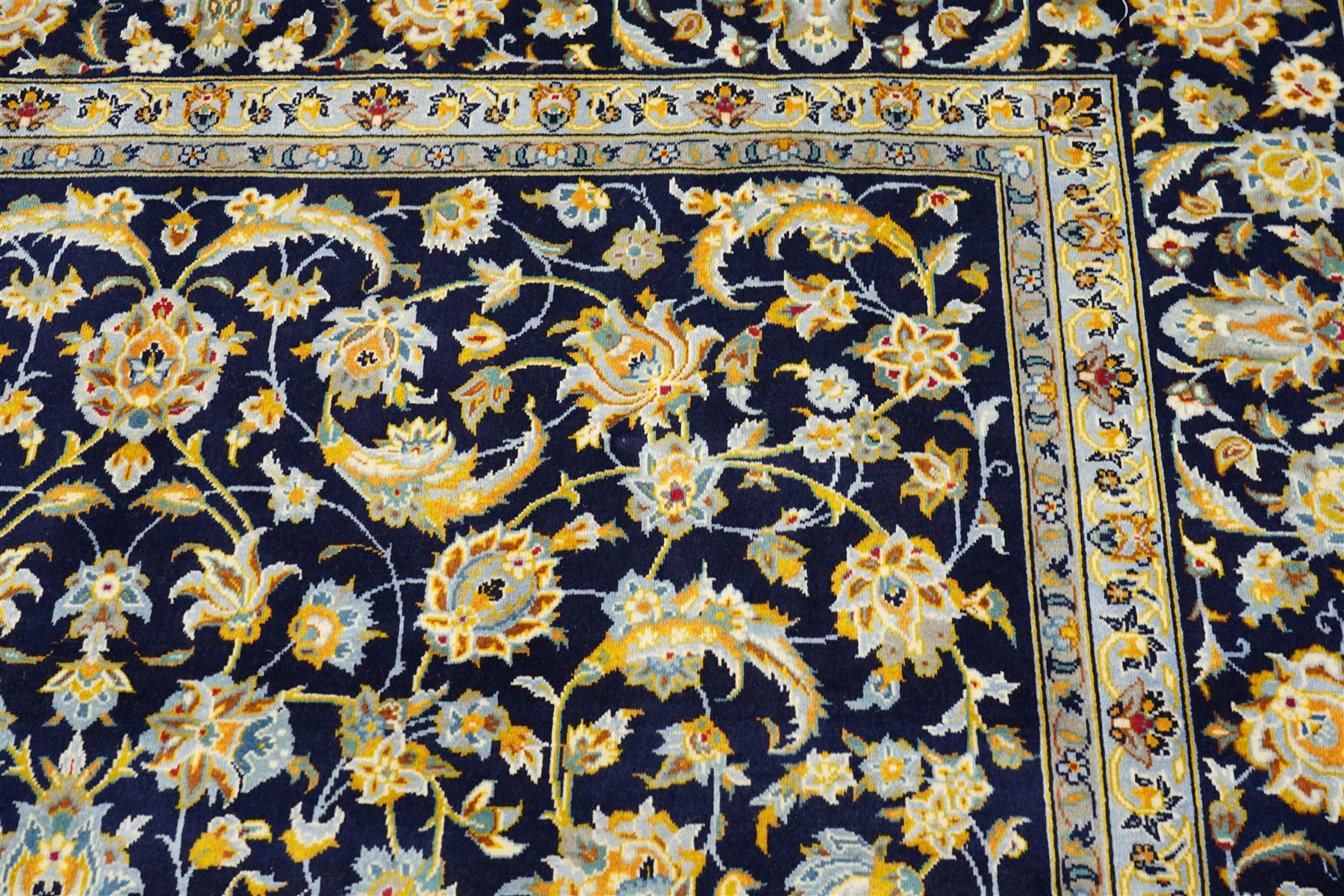 Central Persian Kashan hand-knotted carpet, deep indigo field with a densely all-over arrangement of scrolling floral vines, palmettes, and flowering sprays, in soft ivory, pale blue, and warm gold tones, principal ivory border with a continuous meandering vine of stylized palmettes, and floral motifs, flanked by multiple narrow guard borders, with cotton fringe ends 