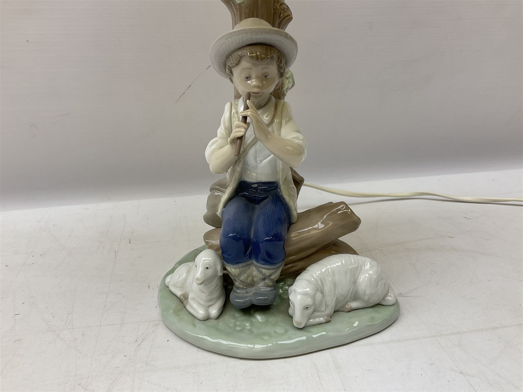 Nao table lamp modelled as a boy playing the flute upon logs with two recumbent sheep at his feet, the stem modelled as a tree trunk, with fabric shade, with printed mark beneath, H46cm