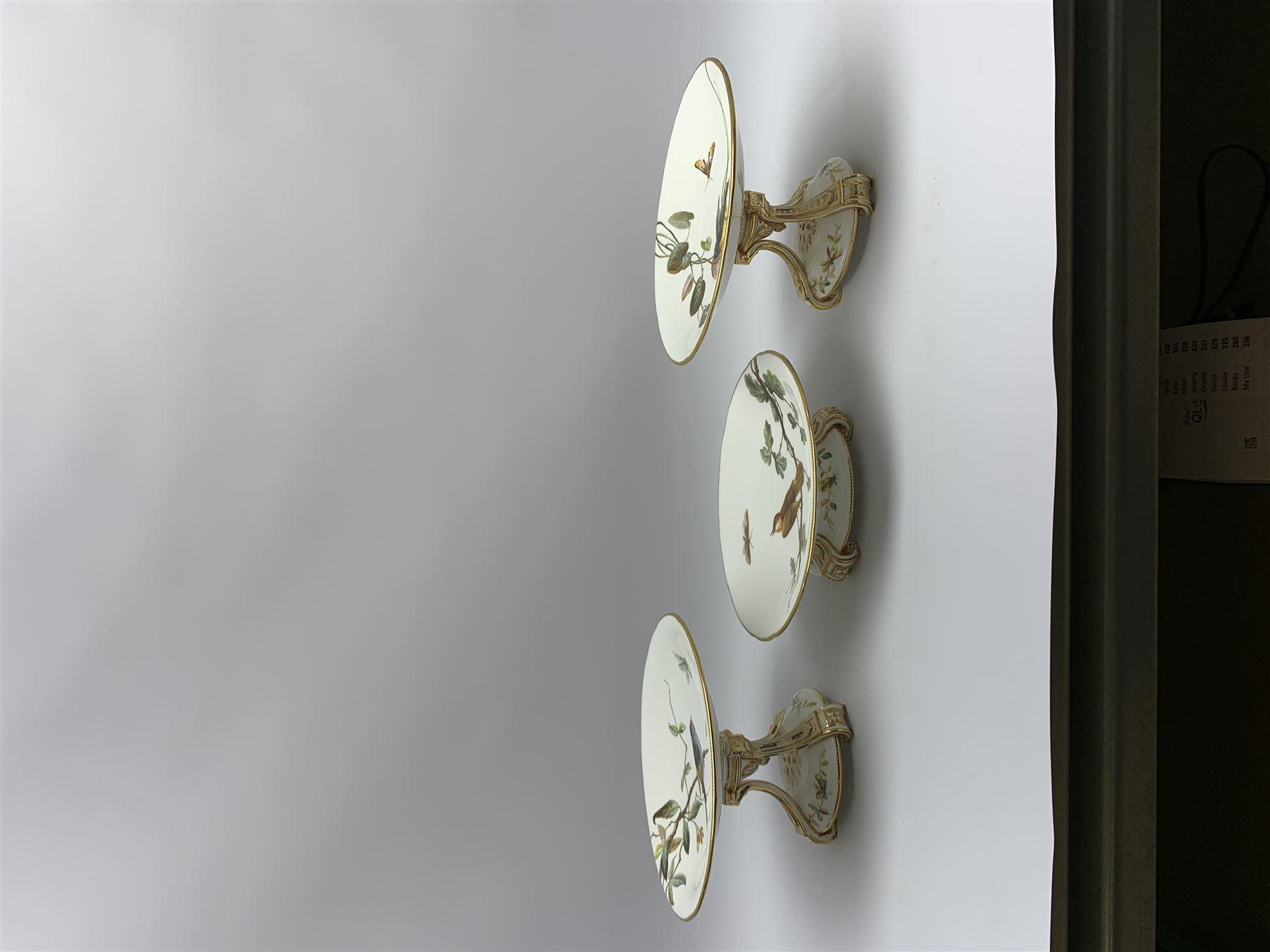 19th century Minton dessert service, comprising four comports, one tazza, and twelve plates, each hand painted with birds perched upon branches, and further detailed with insects and heightened in gilt, with printed retailers mark beneath for John Mortlock Oxford Street London, and impressed Minton mark to most, comport H13.5cm, plates D23.5cm. 
