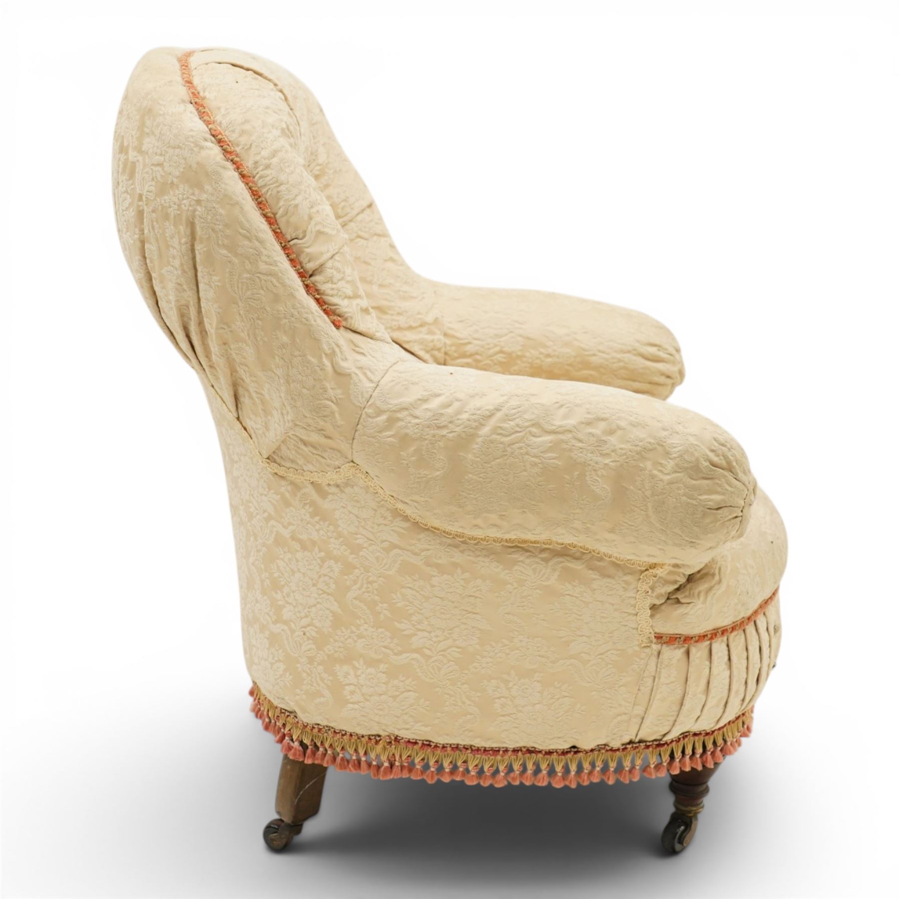 Victorian button-back nursing chair, the rounded horseshoe back enclosing a deep upholstered seat, padded arms and sprung seat in cream floral damask fabric, with crimson piping and tasselled lower fringe, on turned front legs with castors
