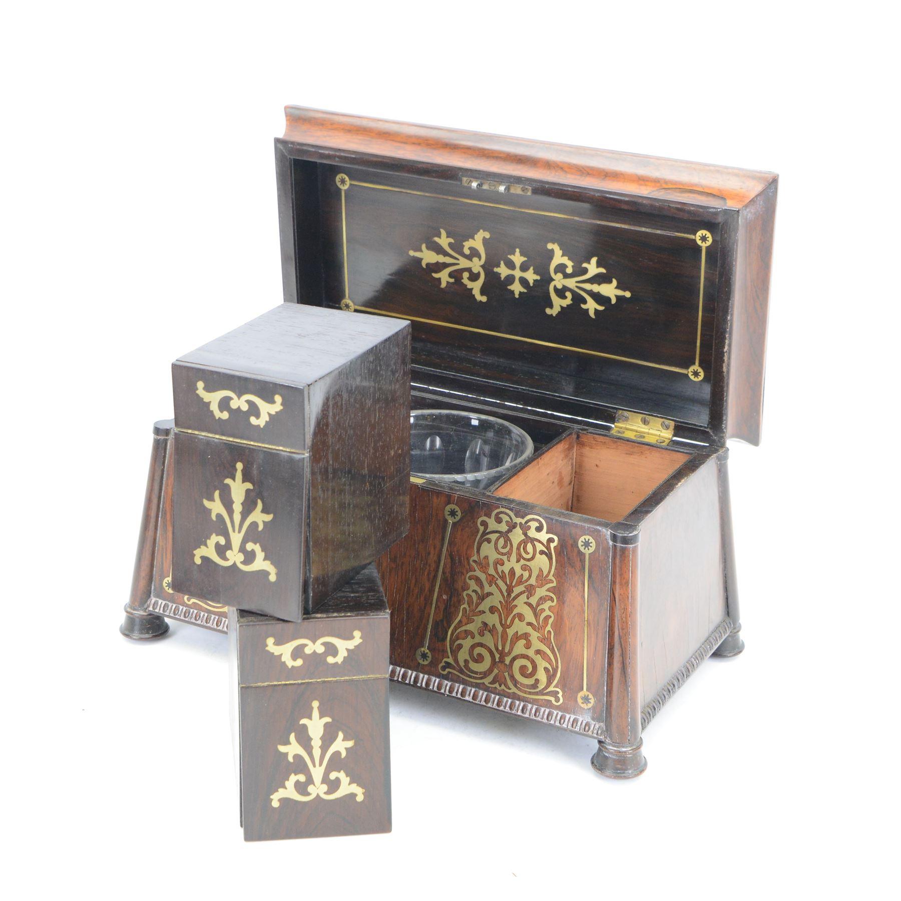 Early 19th century rosewood and brass inlaid tea caddy, of sarcophagus form, the front and hinged opening cover inlaid with brass scrolling panels, opening to reveal a compartmented interior with twin removable boxes with conforming inlaid brass detail to the hinged covers and zinc lining to the interiors, flanking a later glass mixing bowl, the whole upon four turned cylindrical feet, H21cm W36cm D20cm