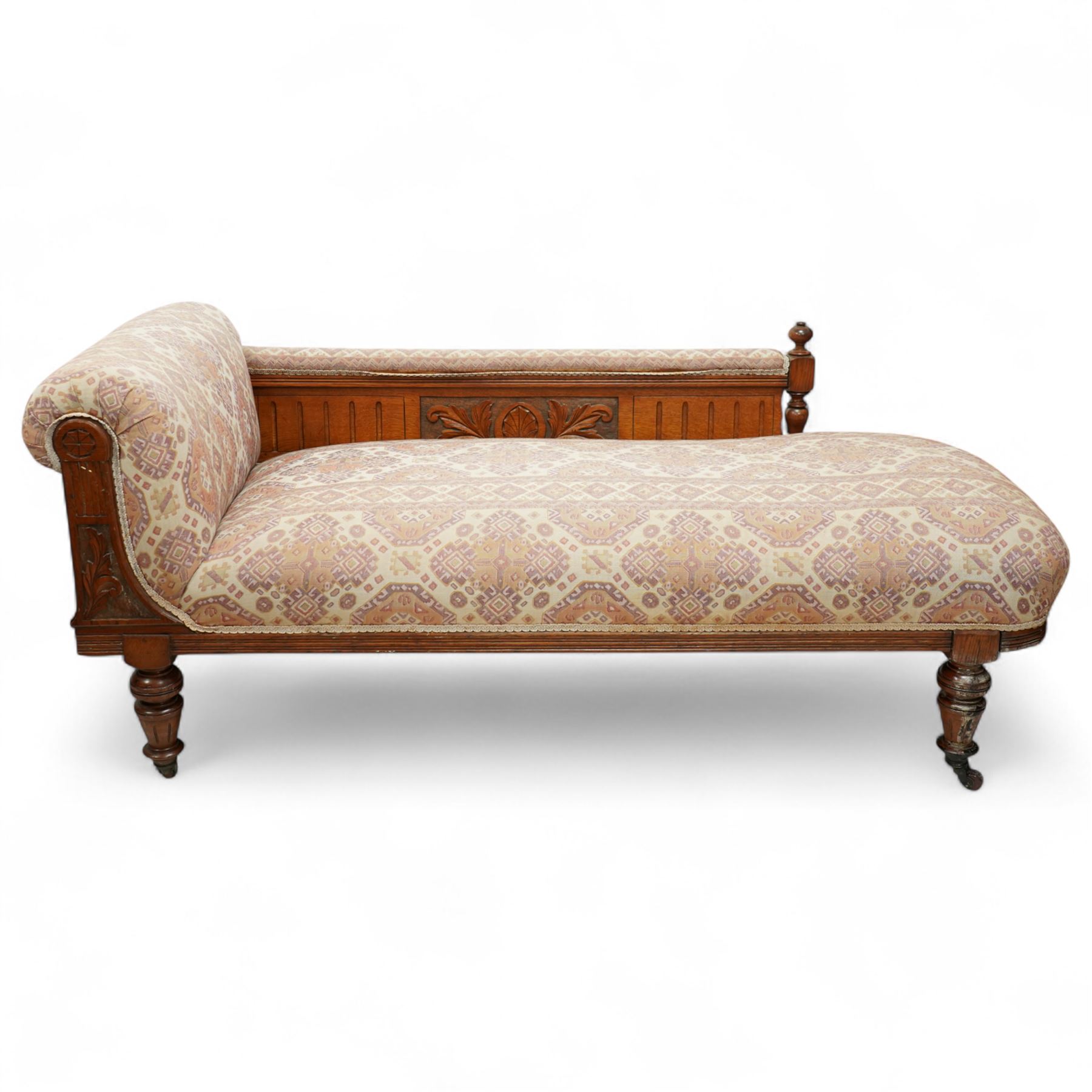 Late Victorian walnut chaise longue, upholstered in geometric patterned fabric, the scroll end and padded back above carved side panel with foliate and roundel motifs, shaped seat rail on turned front supports with ceramic castors