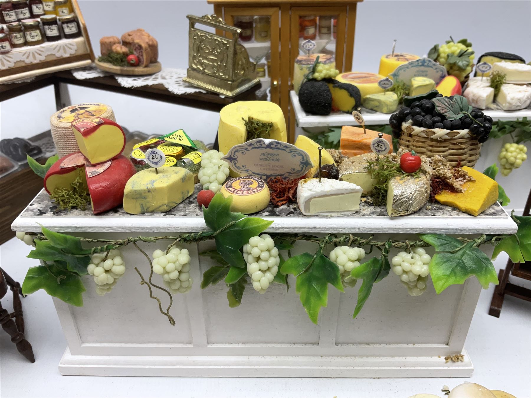 Collection of miniature dolls house grocery shop furniture and produce, comprising glazed corner counter with brass style cash register, shelved preserves unit, pies, cheese board and trays of meats, pair of white cheese counters, biscuit display with six tins, glazed preserves cabinet, pair of shop chairs and four further cheese boards complete with grapes and label flags