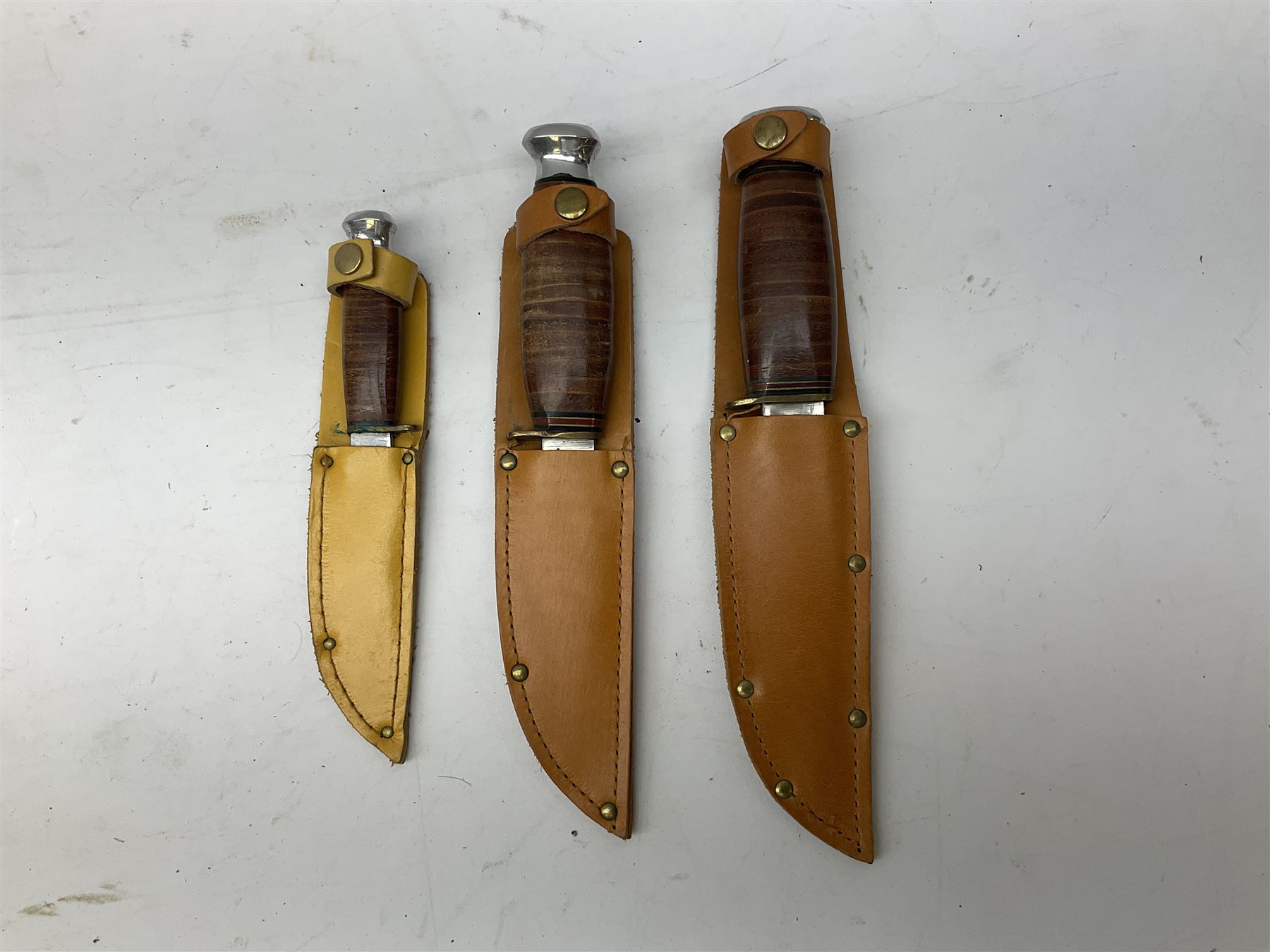 Graduated set of three Bowie knives by J. Nowill & Sons Sheffield England Est. AD1700, each with steel blade, brass cross-piece, stacked leather type grip and alloy pommel; largest blade 13cm and smallest 7.5cm; each in leather sheath (3)