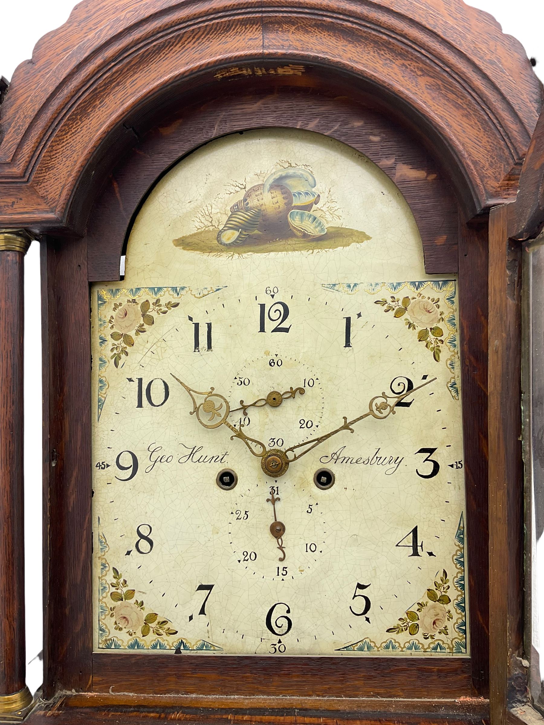 A provincial oak longcase clock retailed by “Geo Hunt, Amesbury” c1820, with a crested break arch pediment and corresponding glazed hood door flanked by two plain pillars with capitals, long trunk with a full length break arch door on a rectangular plinth with a shaped base, painted dial with Arabic numerals, minute markers, subsidiary date and seconds dials and stamped brass hands, matching painted spandrels and a depiction of sea shells to the arch, dial pinned to the movement via a Walker Hughes cast falseplate, with an eight day rack striking movement, striking the hours on a cast bell. With weights and Pendulum




