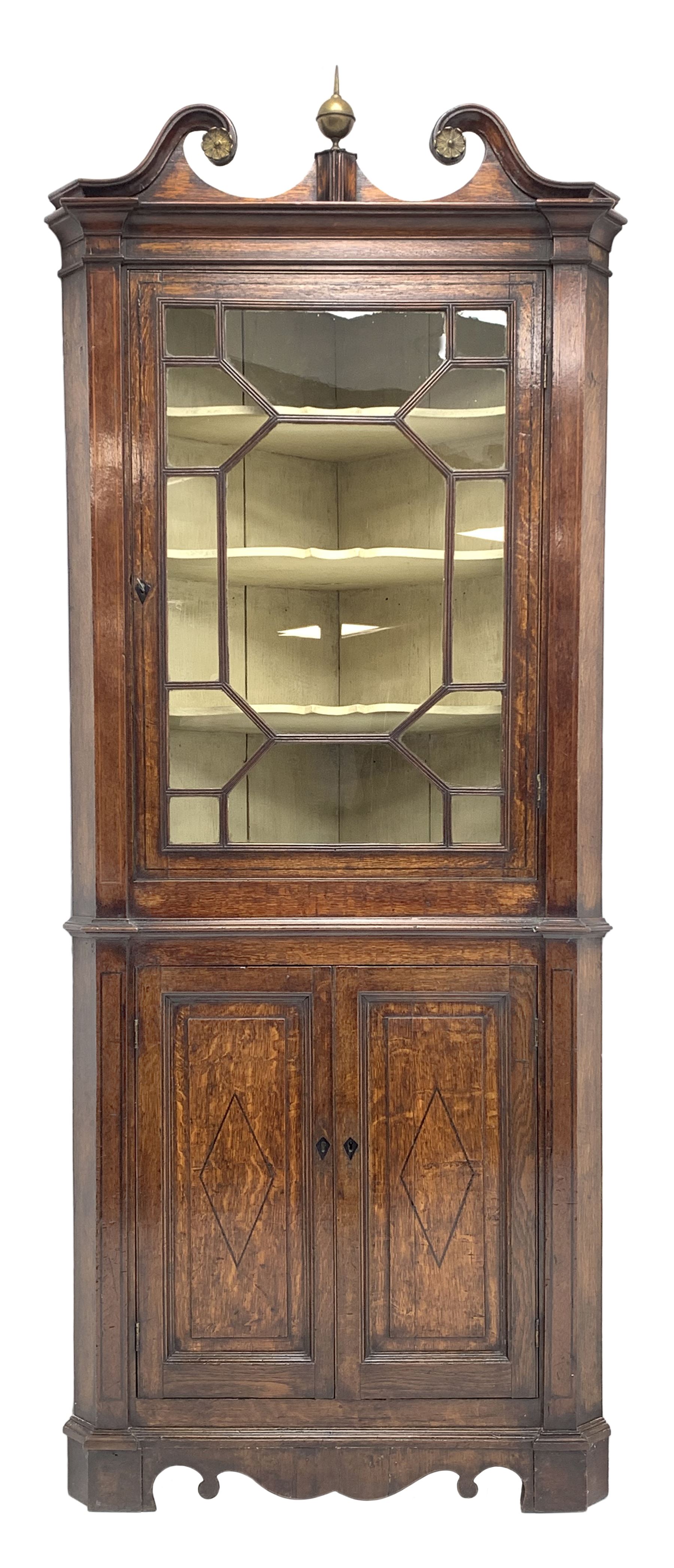 George III oak floor standing corner cabinet, scrolled swan neck pediment with central finial over astragal glazed door, painted interior fitted with three shaped shelves, the lower section fitted with single shaped shelf enclosed by two fielded panel doors inlaid lozenges, on bracket feet, H195cm, W77cm