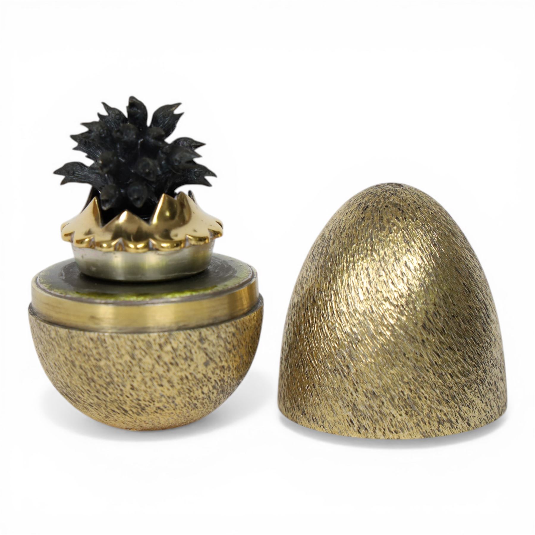 Late 20th century silver and silver-gilt surprise egg, the textured gilt exterior opening to reveal a clutch of chicks, the clustered heads supported upon a spring, within a jagged and lobed silver and gilt encasement, with green enamel surround, hallmarked Stuart Devlin, London 1982, H7cm