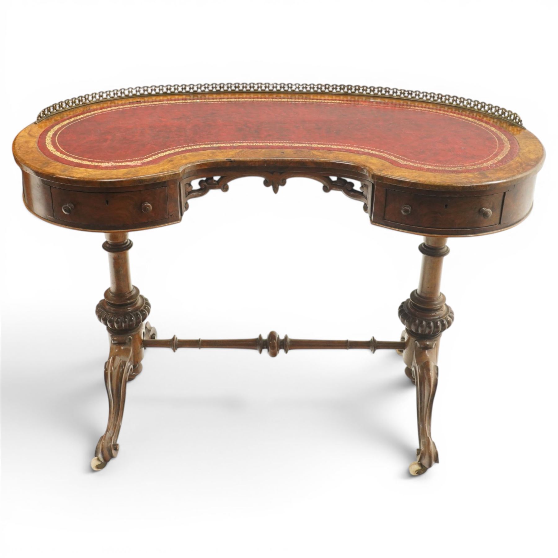 Victorian figured burr walnut kidney-shaped writing table, shaped top with pierced raised bras gallery back over inset red leather writing surface, fitted with two drawers flanking a pierced and carved scrolling apron, raised on turned and gadrooned twin pedestals with scroll-moulded cabriole supports with ceramic castors, united by ring-turned stretcher