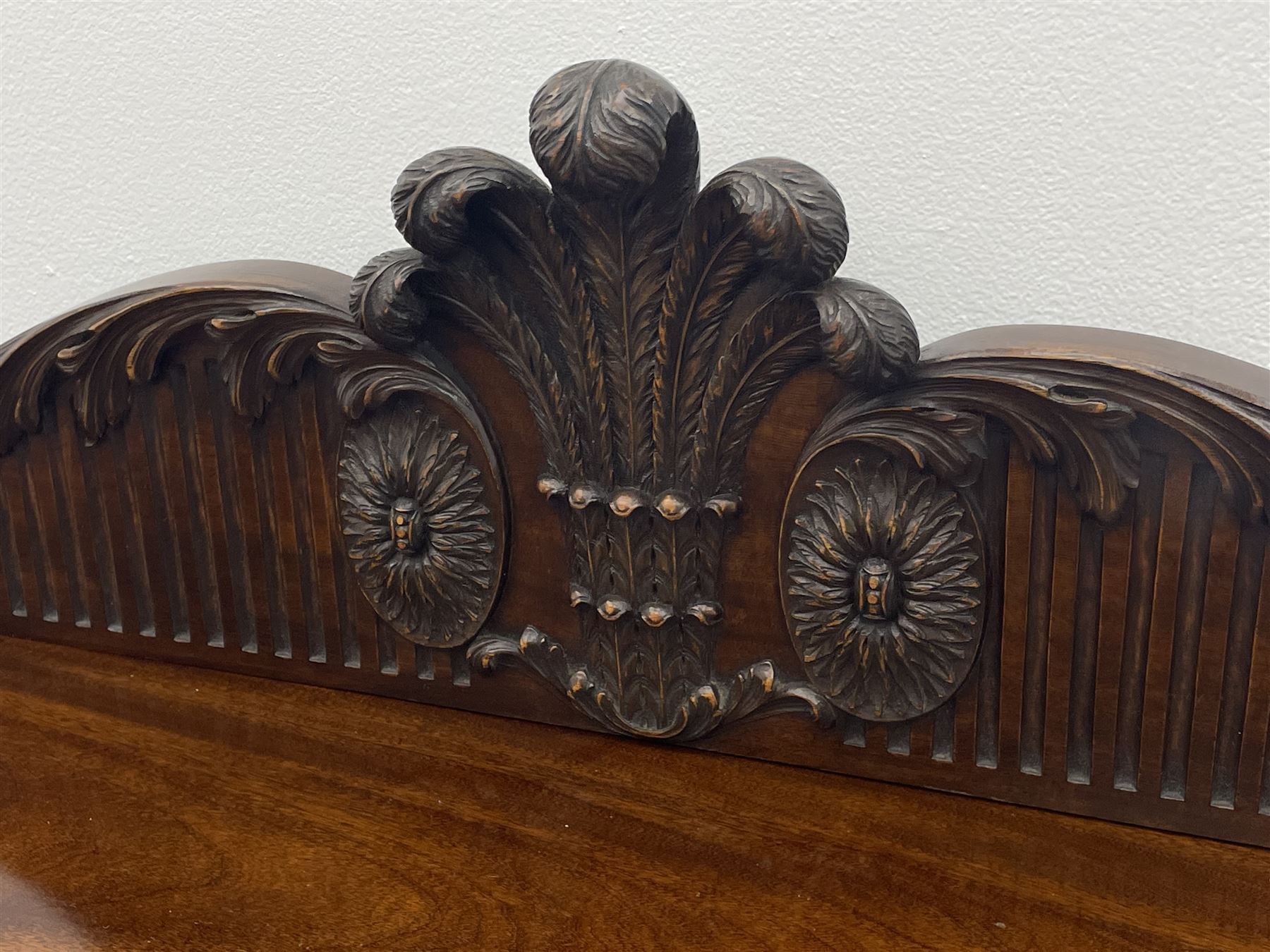 Early 20th century Hepplewhite style serving table, raised fleur-de-lis feather back with fluted detail and flower head mounts, the shaped top with carved foliate edge over single central drawer with applied urn motif, turned and reeded acanthus supports with recessed castors