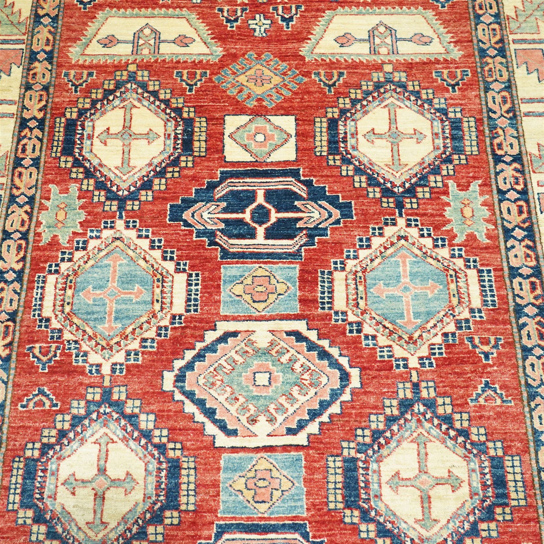Caucasian Kazak hand-knotted rug, madder red field with bold geometric medallions and angular tribal motifs, within multiple patterned borders in a palette of indigo, red, ivory and green 