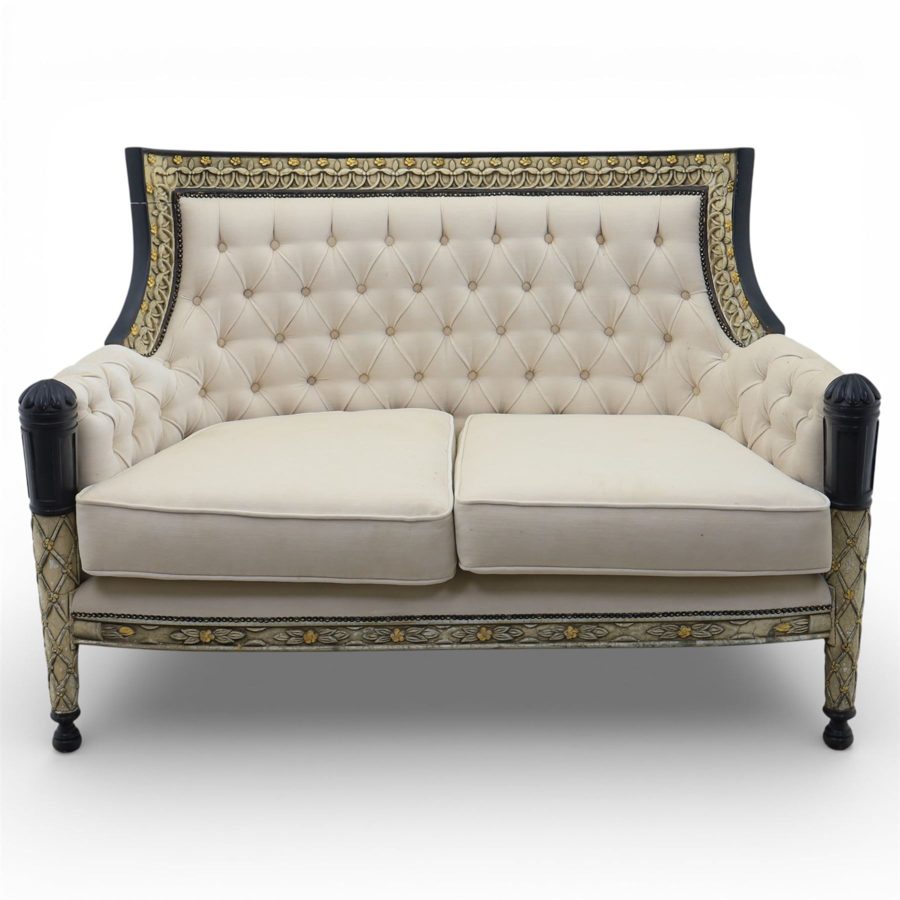 20th century Louis XVI style settee, upholstered in cream fabric with buttoned back and arms, and plain seat cushions, the shaped and black painted frame with entwined foliate detail and gilt flower heads, upon column legs with bun feet