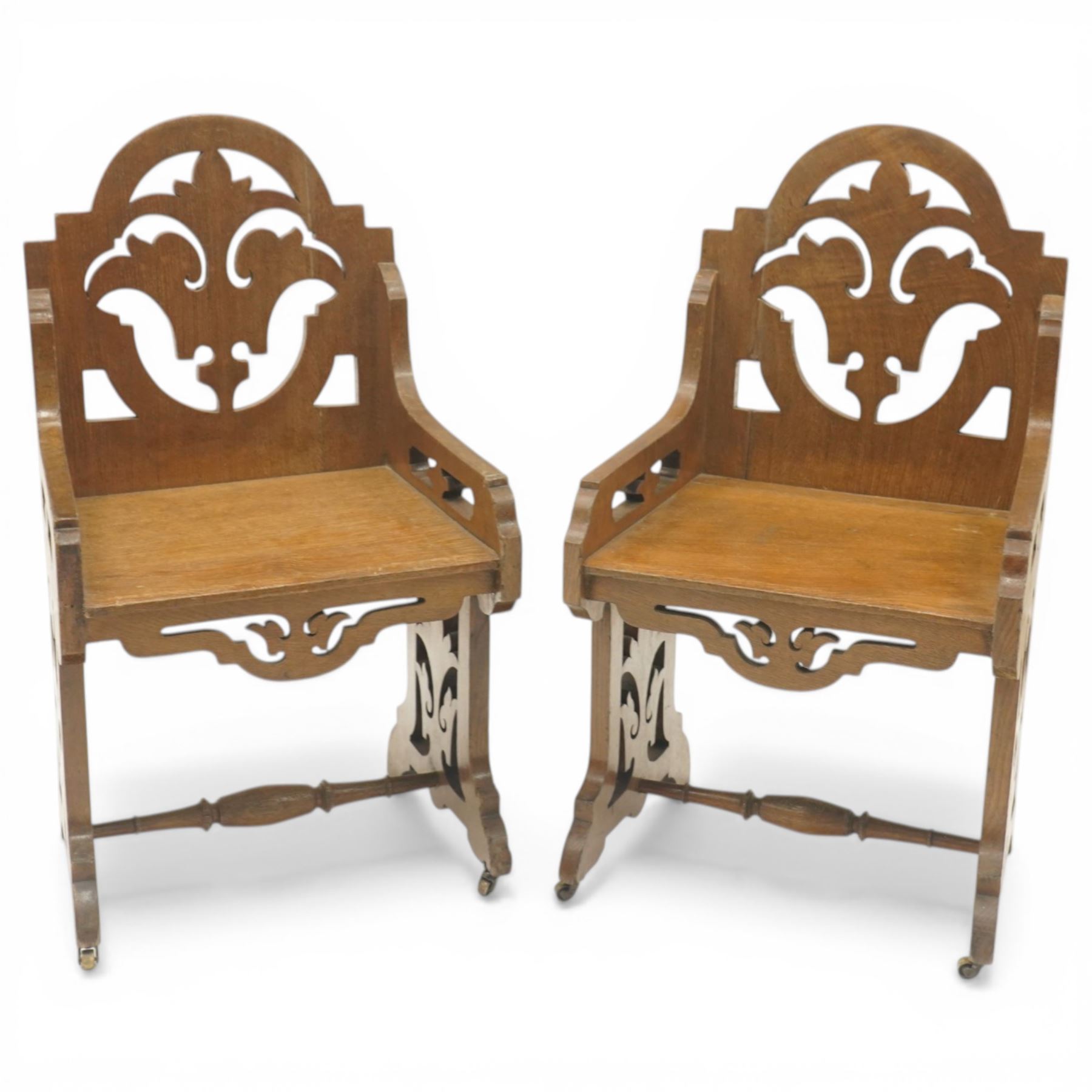 Gothic Revival oak ecclesiastical chairs, shaped cresting rails over pierced foliate splat backs, plank seats above shaped and pierced friezes, raised on shaped end supports united by turned stretchers, on small brass castors