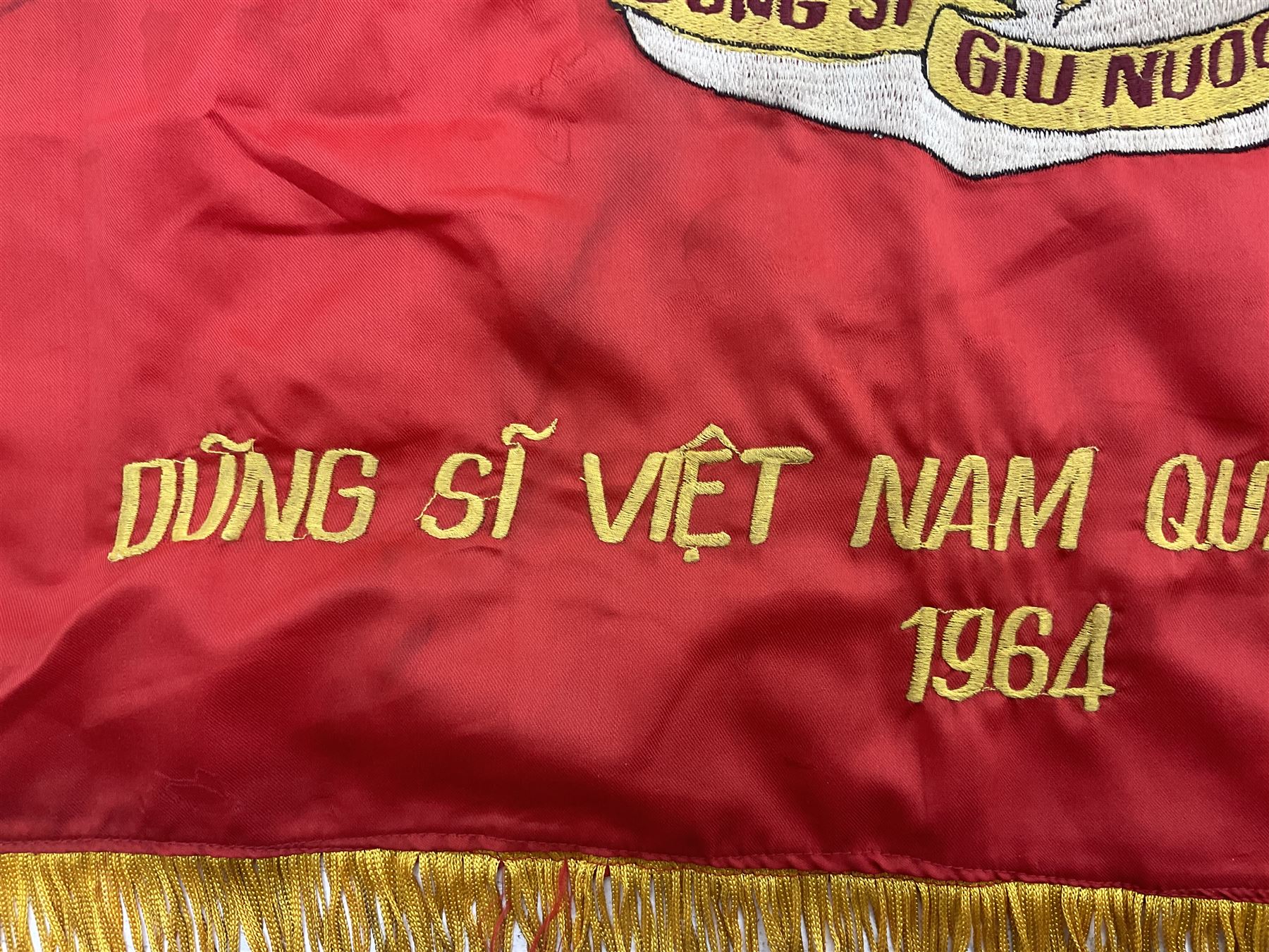1960s North Vietnam banner embroidered in yellow thread on a red ground, roughly translates as 'Peoples Army of Viet Nam. Warriors of Viet Nam Determined To Keep The Country 1964' around a central military crest with the motto 'Warriors Keep The Country'; tassels to three sides 70 x 100cm