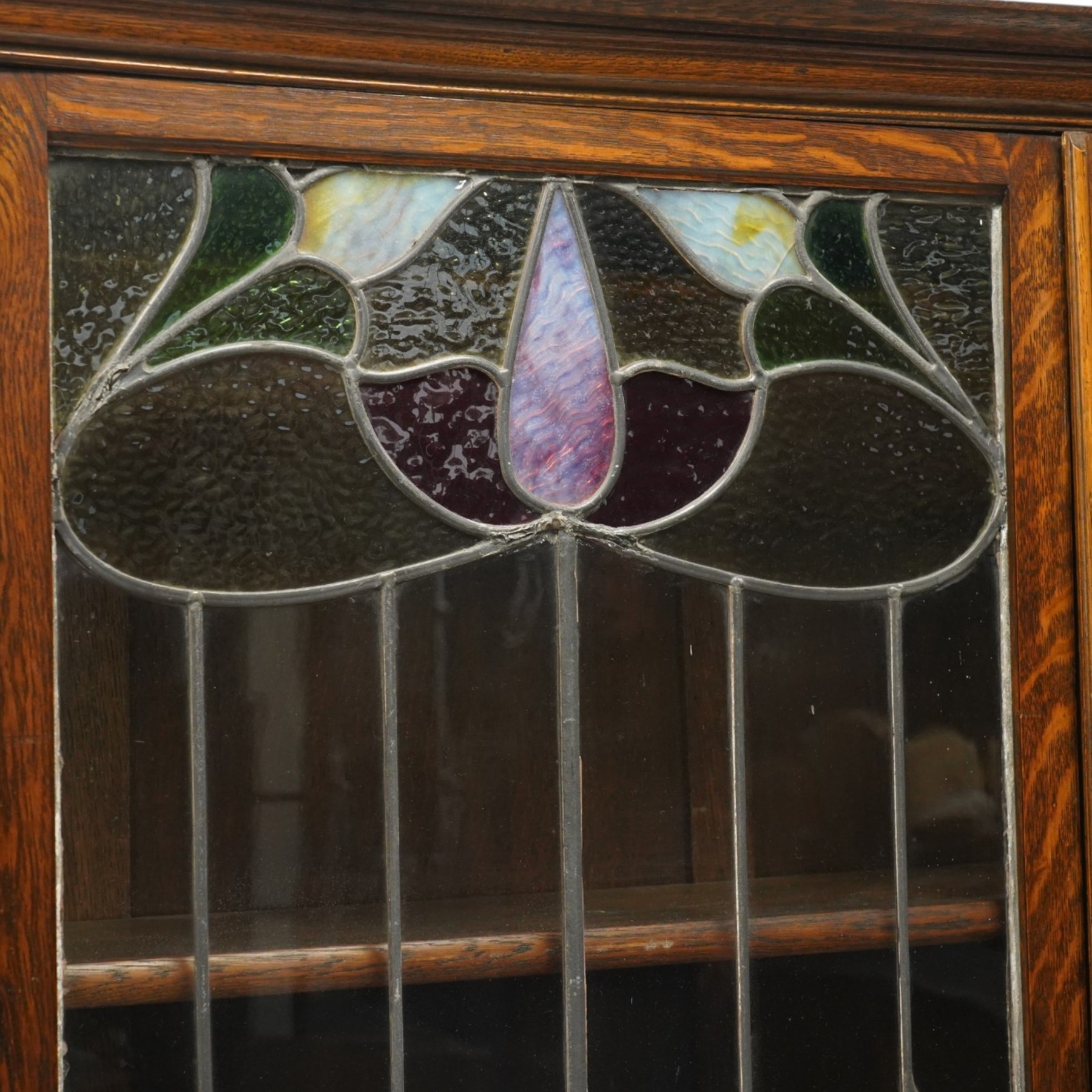 Arts and Crafts oak dresser, projecting moulded cornice above three glazed panels with coloured leaded glass floral panels, flanked by two glazed doors enclosing two adjustable shelves, open mirrored recess below with mirrored back, the base fitted with two short drawers over two central drawers flanked by carved panelled cupboard doors, ornate brass handles, raised on tapering square legs