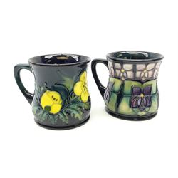 Two Moorcroft mugs of waisted form, the first example decorated in the Violet pattern designed by Sally Tuffin, the second example decorated in the Buttercup pattern designed by Sally Tuffin, each with impressed marks beneath, H8.5cm. 