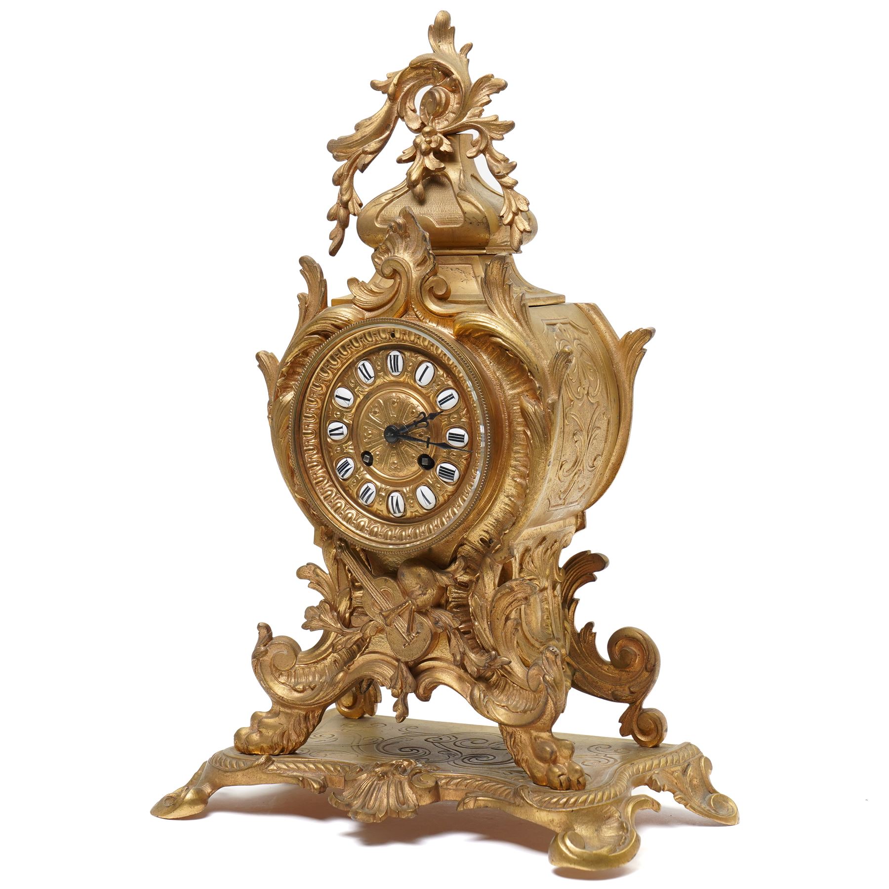 French-Late 19th century 8-day gilt brass rococo style mantel clock, c1890, waisted case  decorated with scroll work, leaves and musical instruments, resting on an attached plinth with splayed feet, dial within a glazed brass bezel with a gilt repoussé centre, porcelain cartouche Roman numerals and steel fleur-di-Lis hands, with a twin train movement, recoil anchor escapement and countwheel strike, sounding the hours and half-hours on a bell. With a matching pair of five-light candelabra. No Pendulum or Key.