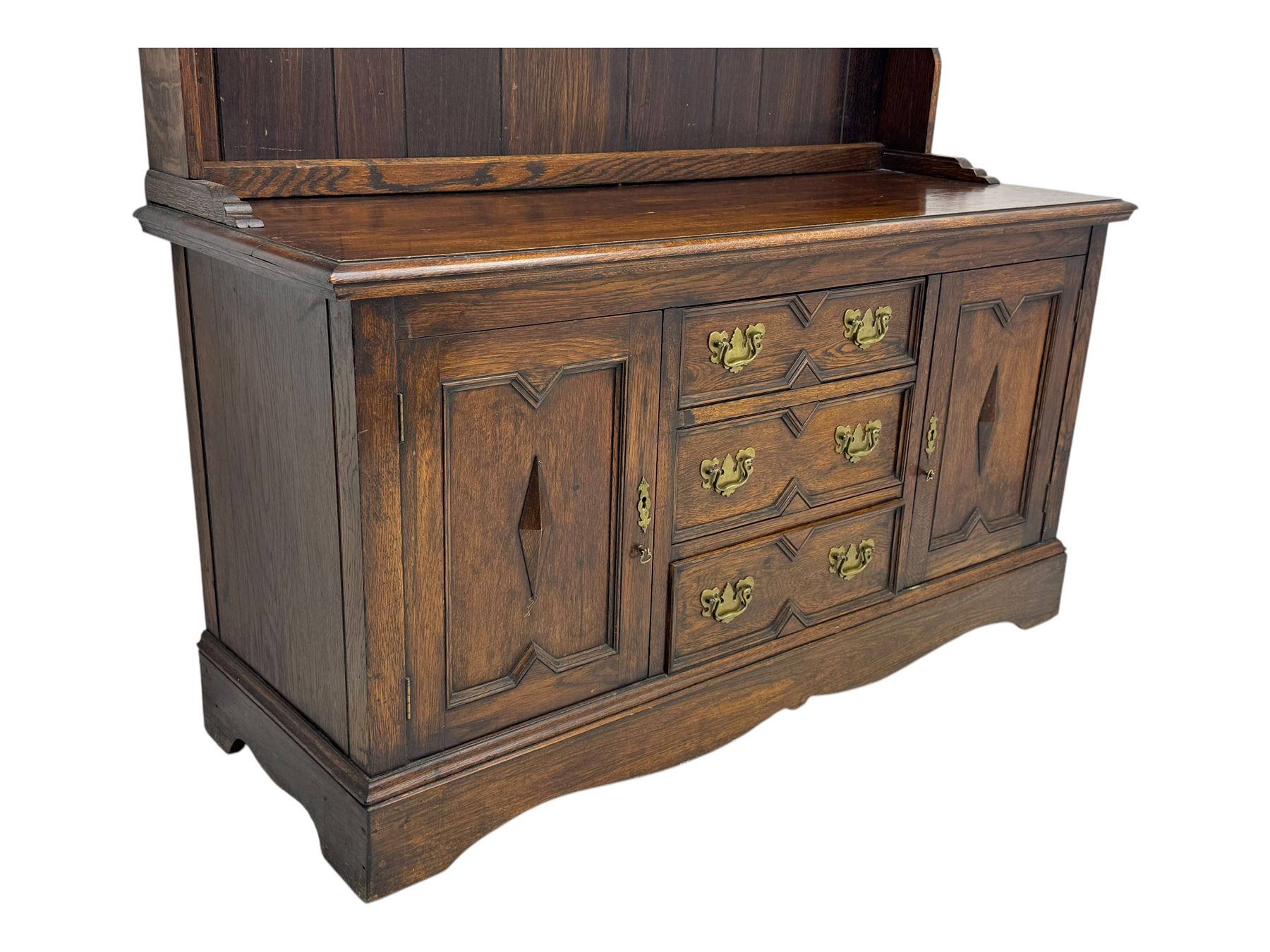 Late 19th century oak dresser, the upper section with two shelves beneath a shaped cornice, the lower section with a central bank of three drawers flanked by cupboard doors, all decorated with geometric diamond motifs, the drawers fitted with brass handles, raised on a shaped plinth base