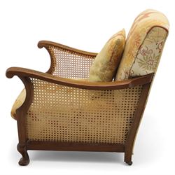 Pair of mid-20th century mahogany framed bergère armchairs, each with carved scroll arms and caned side panels, floral upholstered loose cushions, raised on cabriole front supports