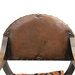 Early 20th century mahogany Savonarola chair, arched leather back with brass studwork, curved open arms with incised geometric decoration, loose patchwork velvet cushion seat, X-frame supports united by circular side rails, on scrolled feet