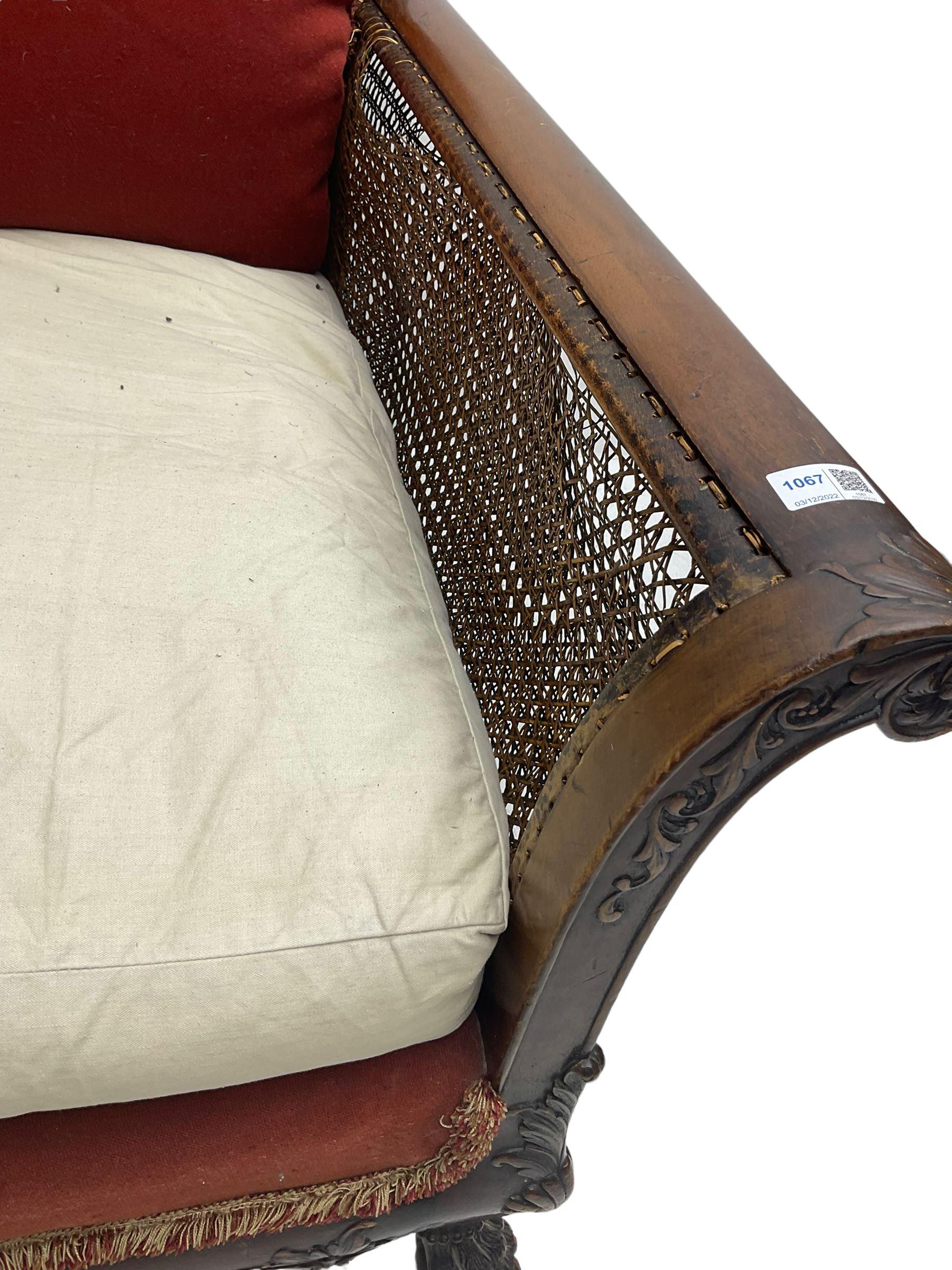 Early 20th century bergère three seat settee, shaped shell carved cresting rail, double caned back and sides, scrolled upright arms and shaped apron carved with shell and foliage, on hairy paw carved feet