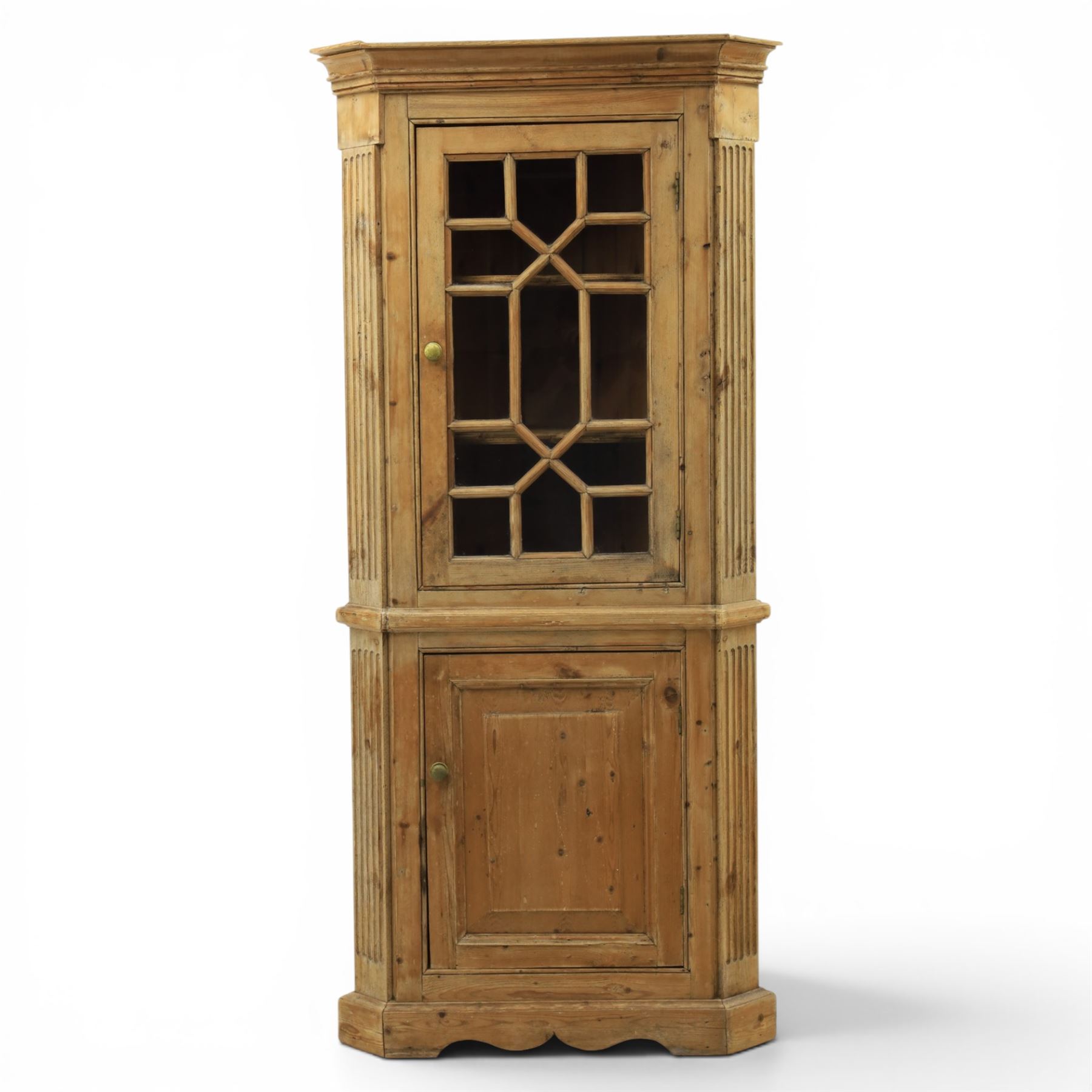 19th century stripped pine corner cabinet, projecting cornice flanked by fluted sides, astragal glazed upper door with brass knob, lower section with fielded panelled door, raised on a shaped plinth base 