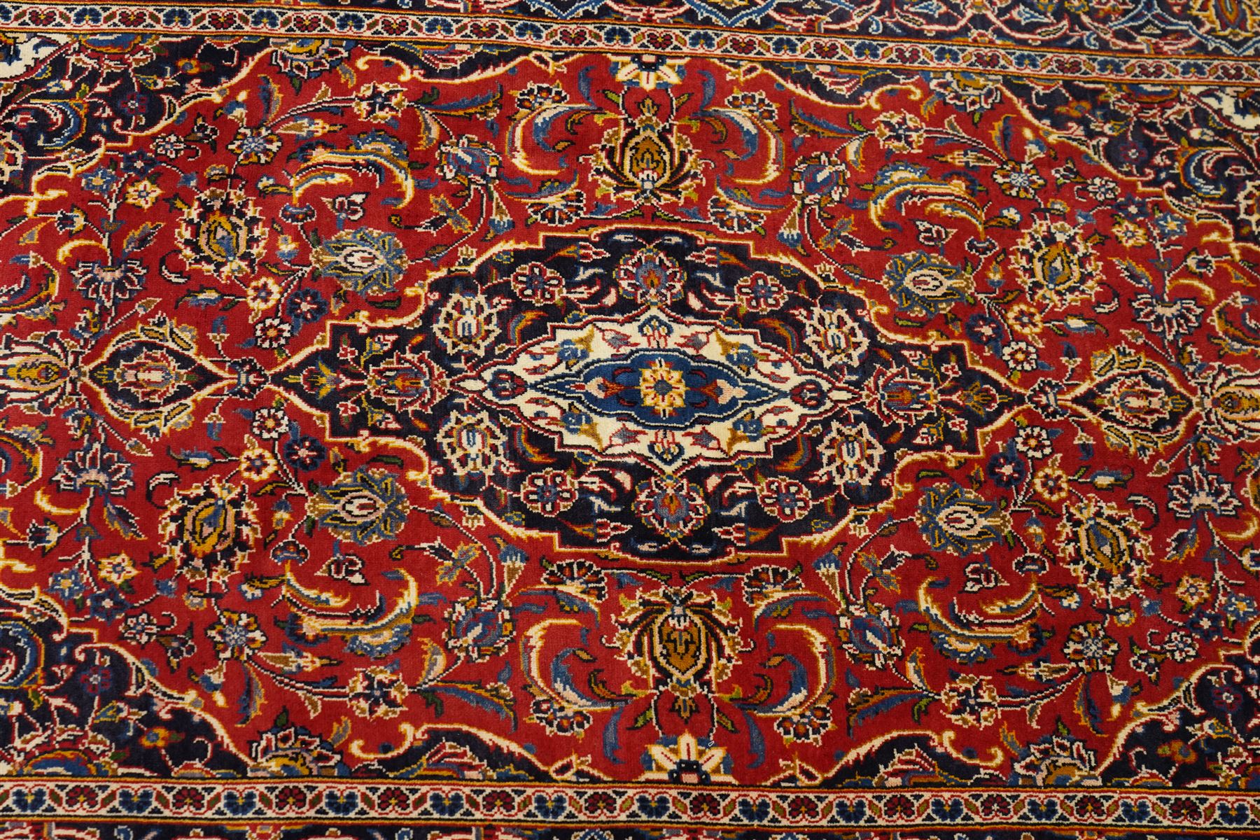 Central Persian Kashan hand-knotted rug, madder red field, with central lobed medallion with pendants, issuing an all-over design of scrolling vine, palmettes, and floral sprays, with conforming spandrels, enclosed by a broad indigo palmette and vine border framed by guard stripes in a palette of navy, blue and ivory tones, with cotton fringe ends 