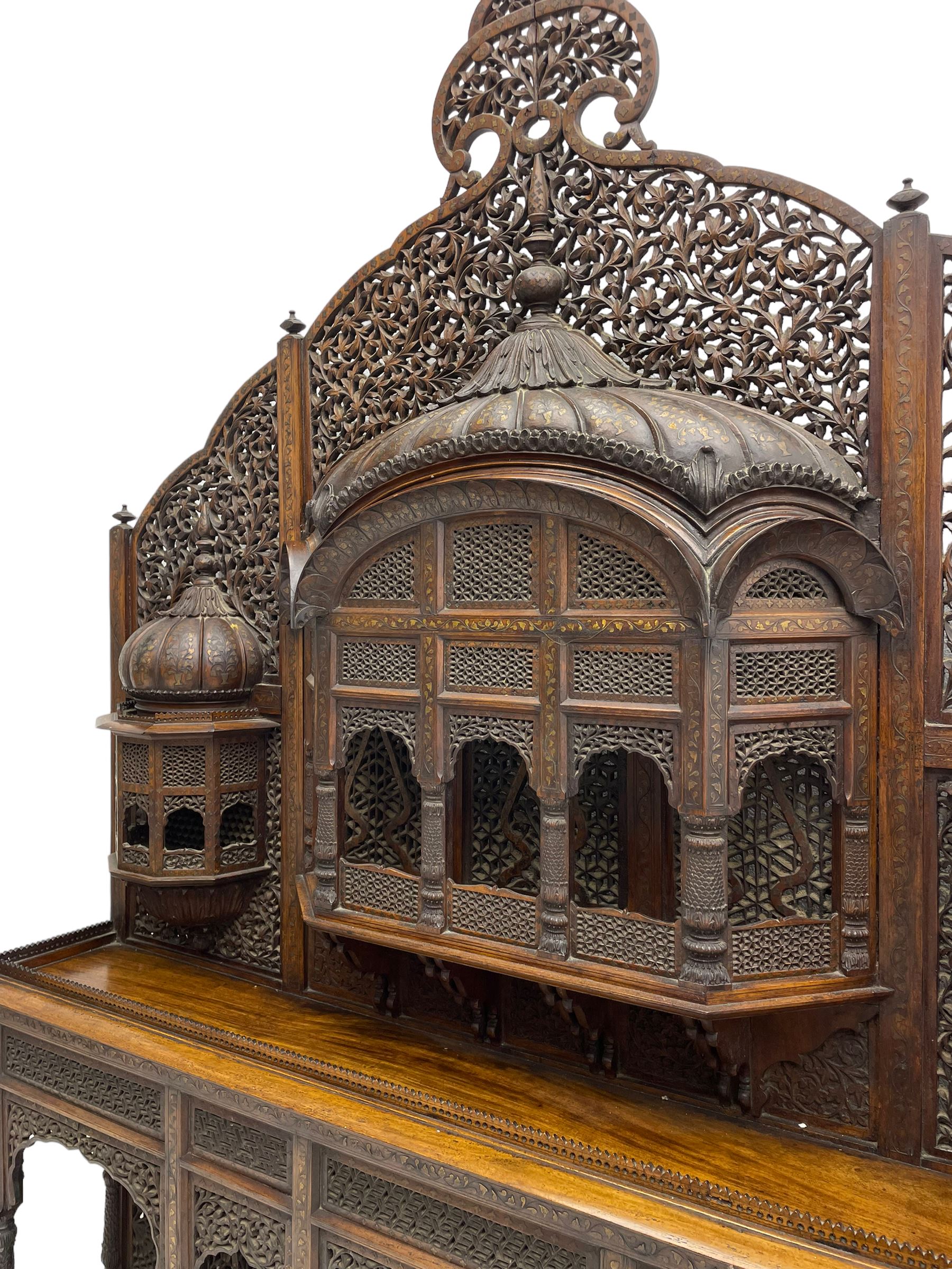 Late 19th century Indian Burmese carved and pierced hardwood and brass inlaid wall shelf, the shaped back profusely decorated with trailing and interlaced foliage, with projecting architectural structures, the central balcony with canopy top and five open windows with arched apertures on columns, two flanking five-sided balconies, the shelf supported by six foliate carved columns with pierced cusped pointed arches 