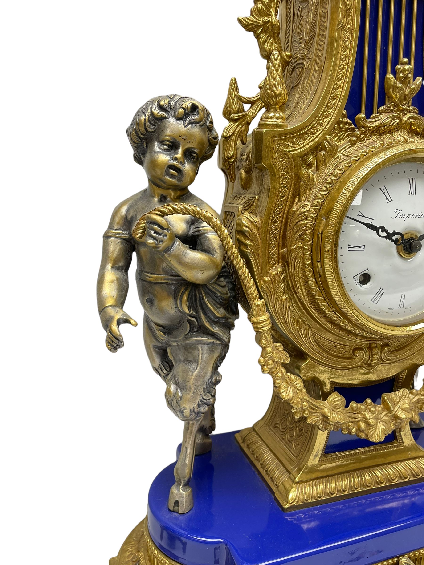 A 20th century continental gilt metal Lyre mantle clock with a pair of matching six light candelabra, spring driven movement housed in a gilt drum case on a brass and cobalt blue base with paw feet, two bronzed metal figures of mythological cherubs with animal legs and cloven hooves supporting a festooned garland swag, eight-day twin barrel striking movement with a floating lever balance escapement, striking the hours and half-hours on two bells, white enamel dial with roman numerals, minute track and pierced steel hands, dial inscribed “Imperial”. 