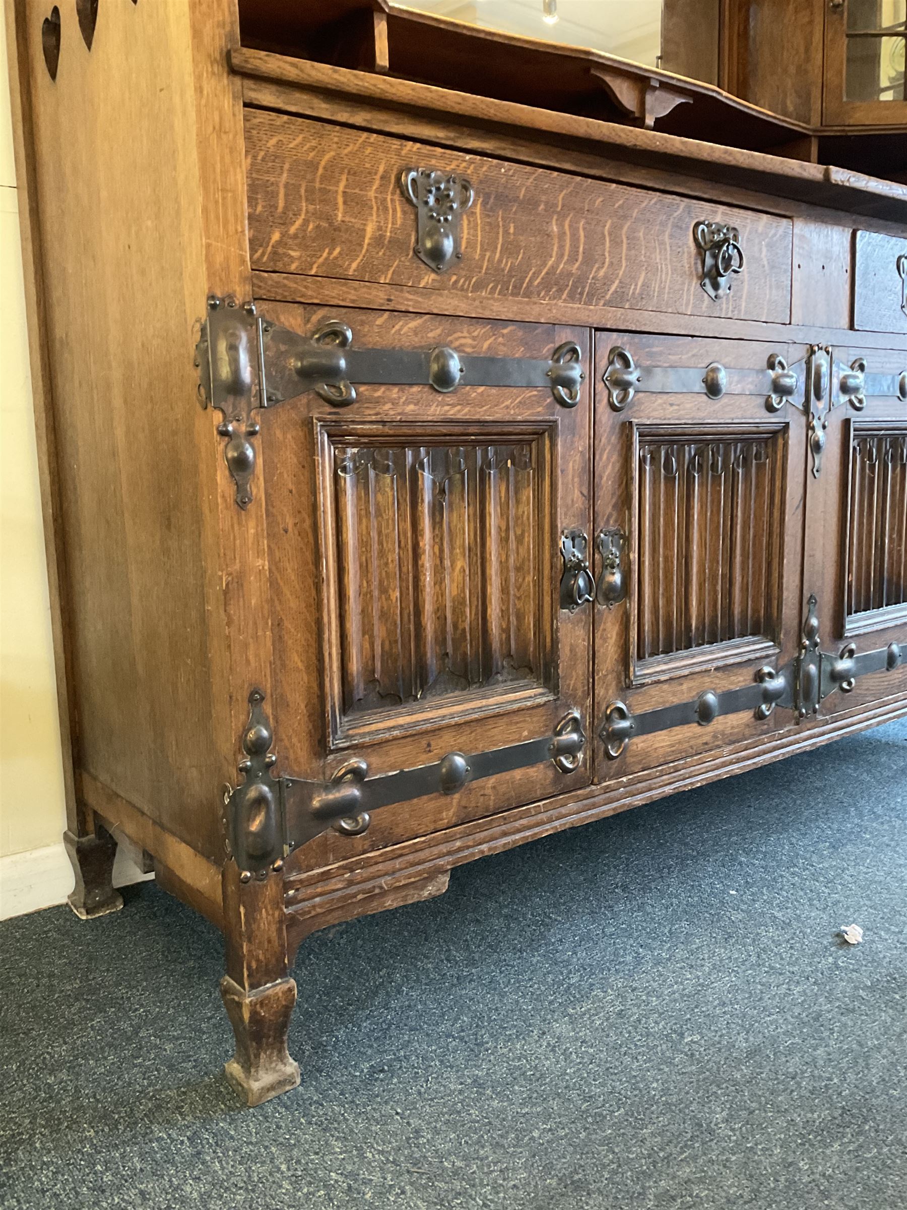 Shapland and Petter - Arts & Crafts period oak dresser, raised gallery top with spade balustrade, central bevelled mirror flanked by two cabinet, astragal glazed doors with yellow tinted glass and bullseye panes, the sides with pierced heart shaped decoration, fitted with two drawers and two double cupboards, enclosed by linenfold panelled doors,  copper work strap hinges and fixtures, on shaped spade feet, the locks stamped 'S&P'