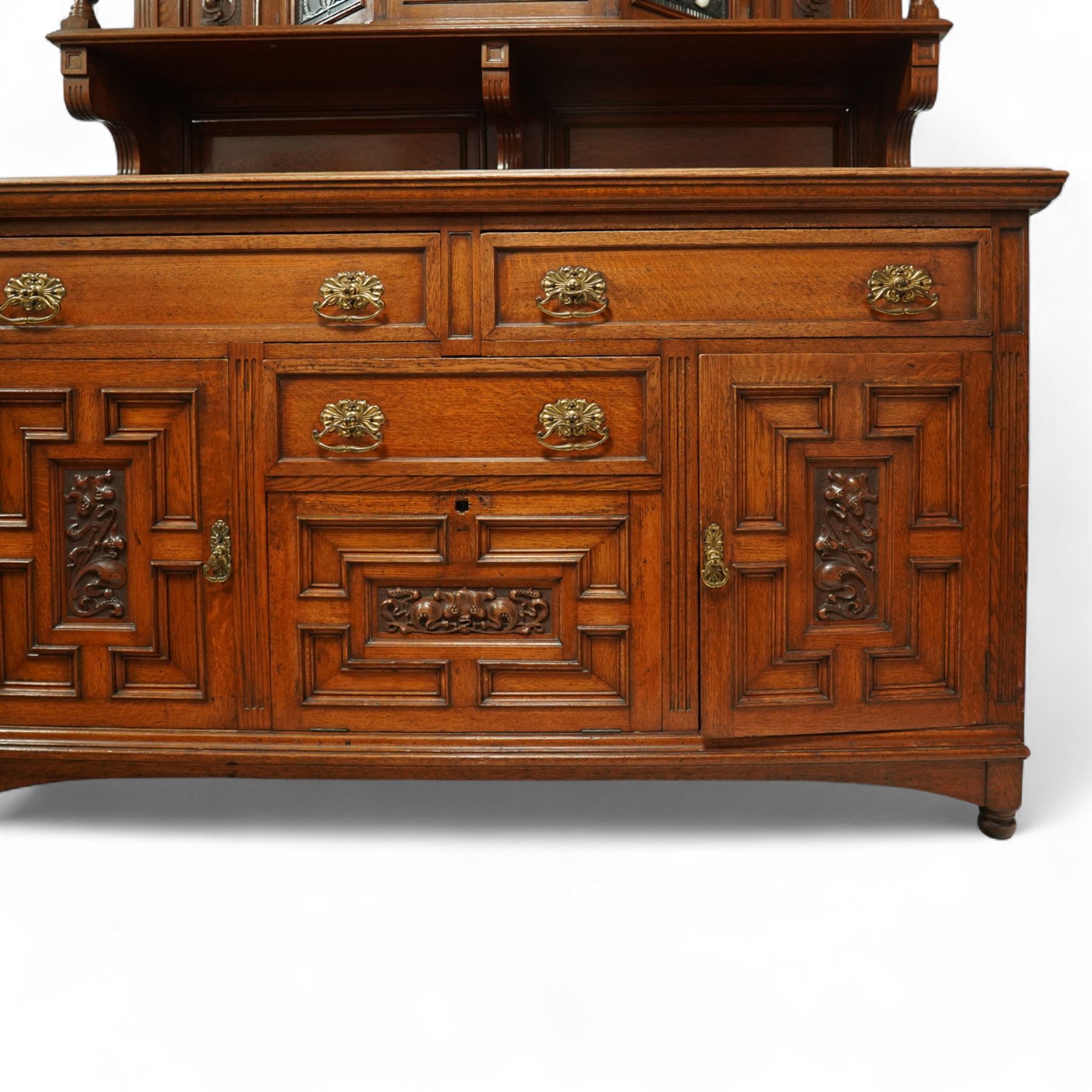 Early 20th century golden oak dresser sideboard, superstructure with three stained and leaded glass doors flanked by carved uprights above an open display shelf, the base with three drawers and geometric panelled cupboard doors, enclosing drawers with cutlery and bottle compartments, raised on short feet