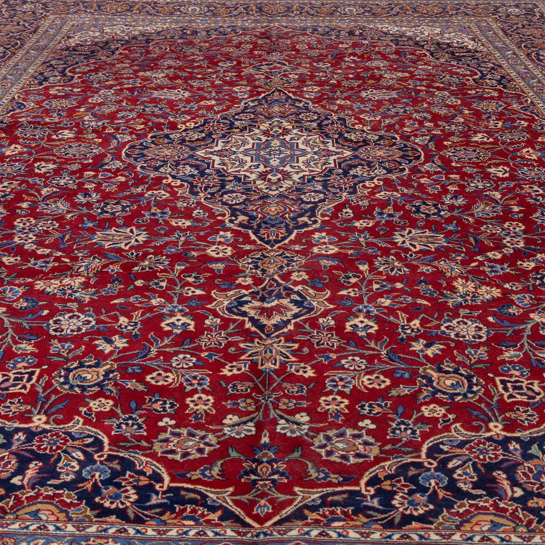 Central Persian Kashan hand-knotted carpet, madder red field with central cusped medallion in navy and ivory enclosing a floral rosette and radiating scrolling arabesques and palmettes, across a densely ornamented ground of finely drawn vinery enriched with a profusion of flowering sprays and stylized blossoms, spandrels in deep indigo and principal border of navy ground decorated with a continuous palmette and vine-scroll flanked by multiple guard stripes, with cotton fringe ends